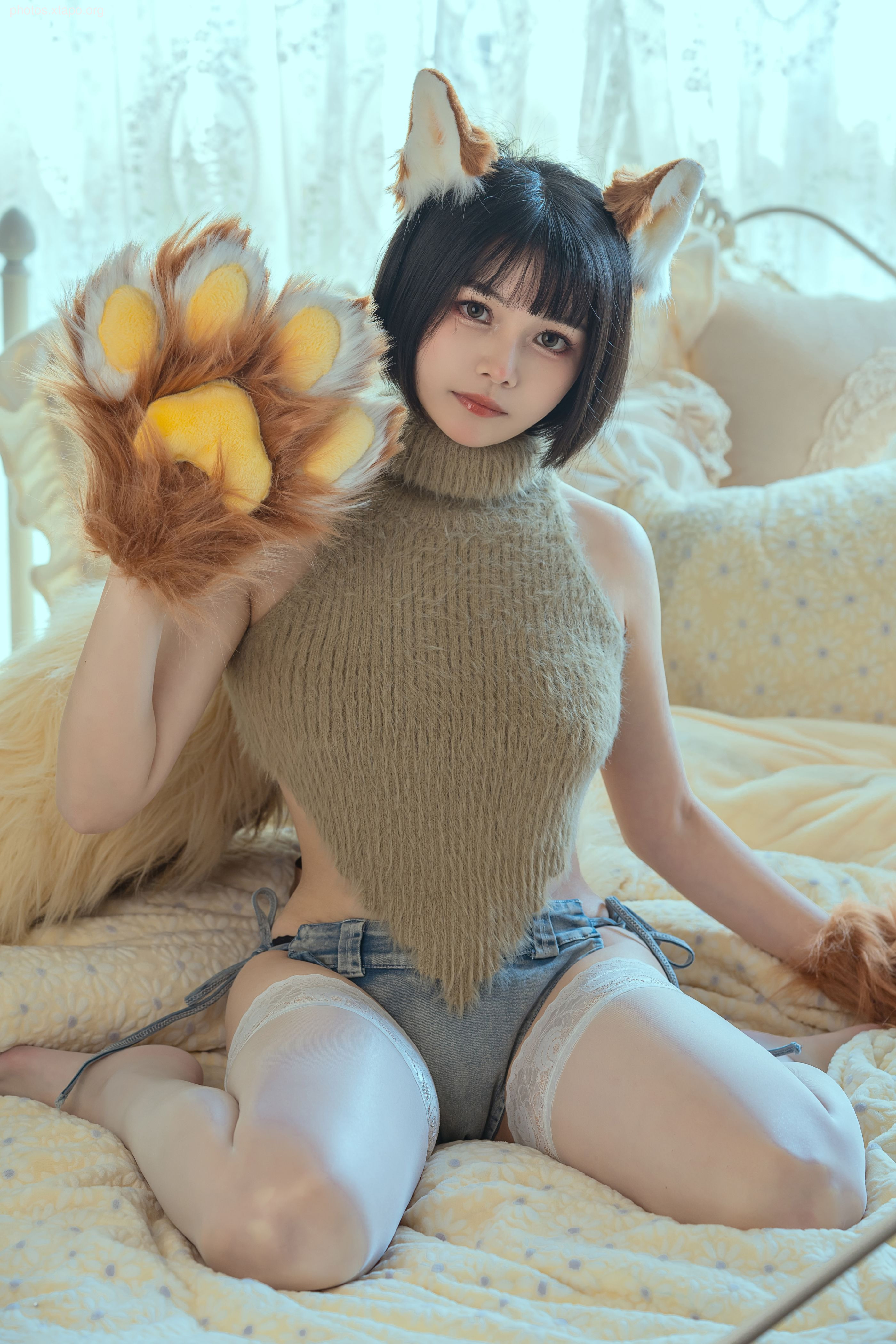 A young woman with short black hair sits on a bed, wearing a light brown knit sweater, denim shorts, and white thigh-high stockings. She holds a fluffy brown paw.  She's wearing cat ears headband.