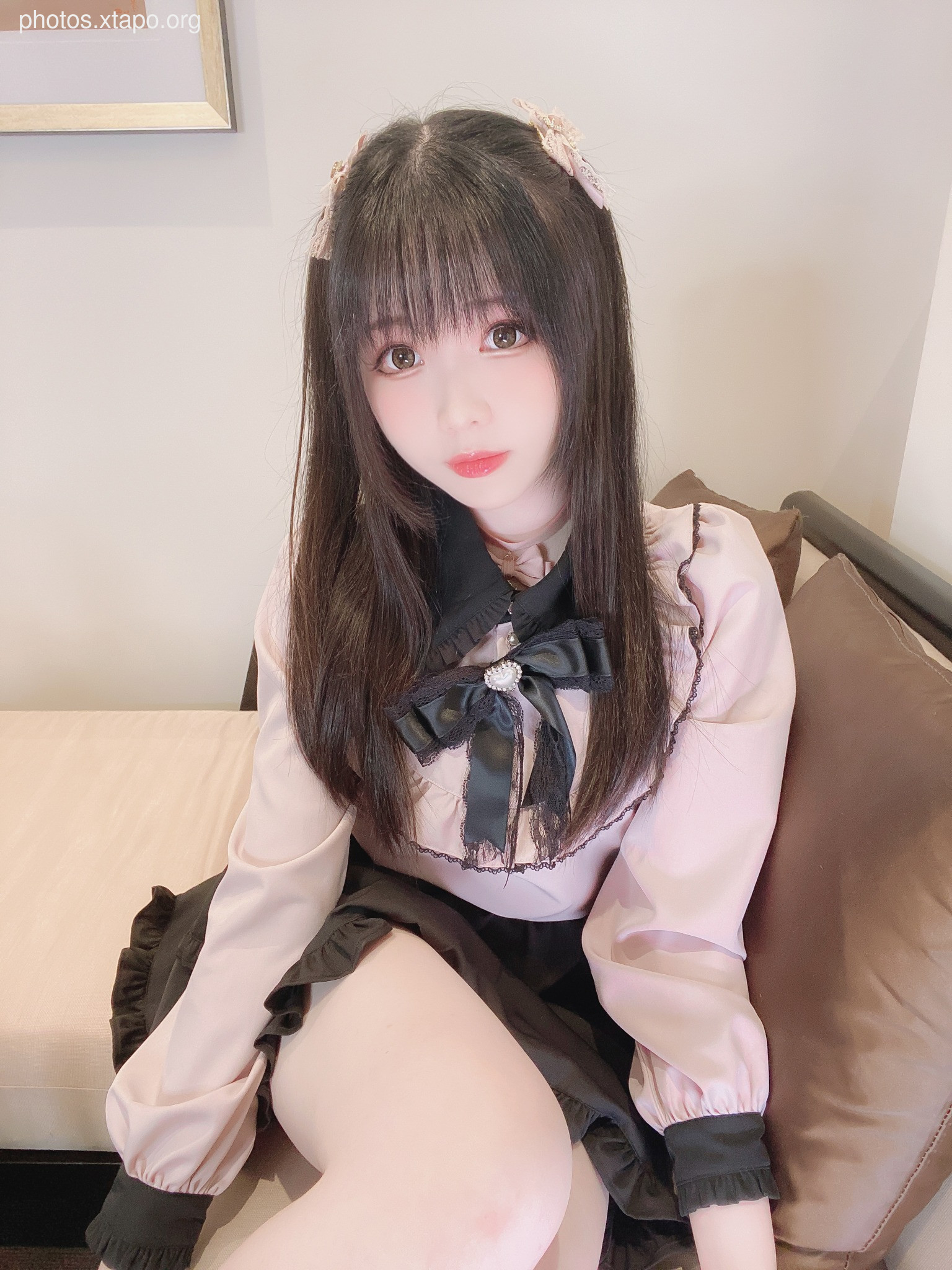 A young woman with long dark hair and bangs poses in a pink ruffled blouse and black skirt. She is wearing small hair clips and has a bow tie on her blouse. The photo was taken indoors against a neutral background.