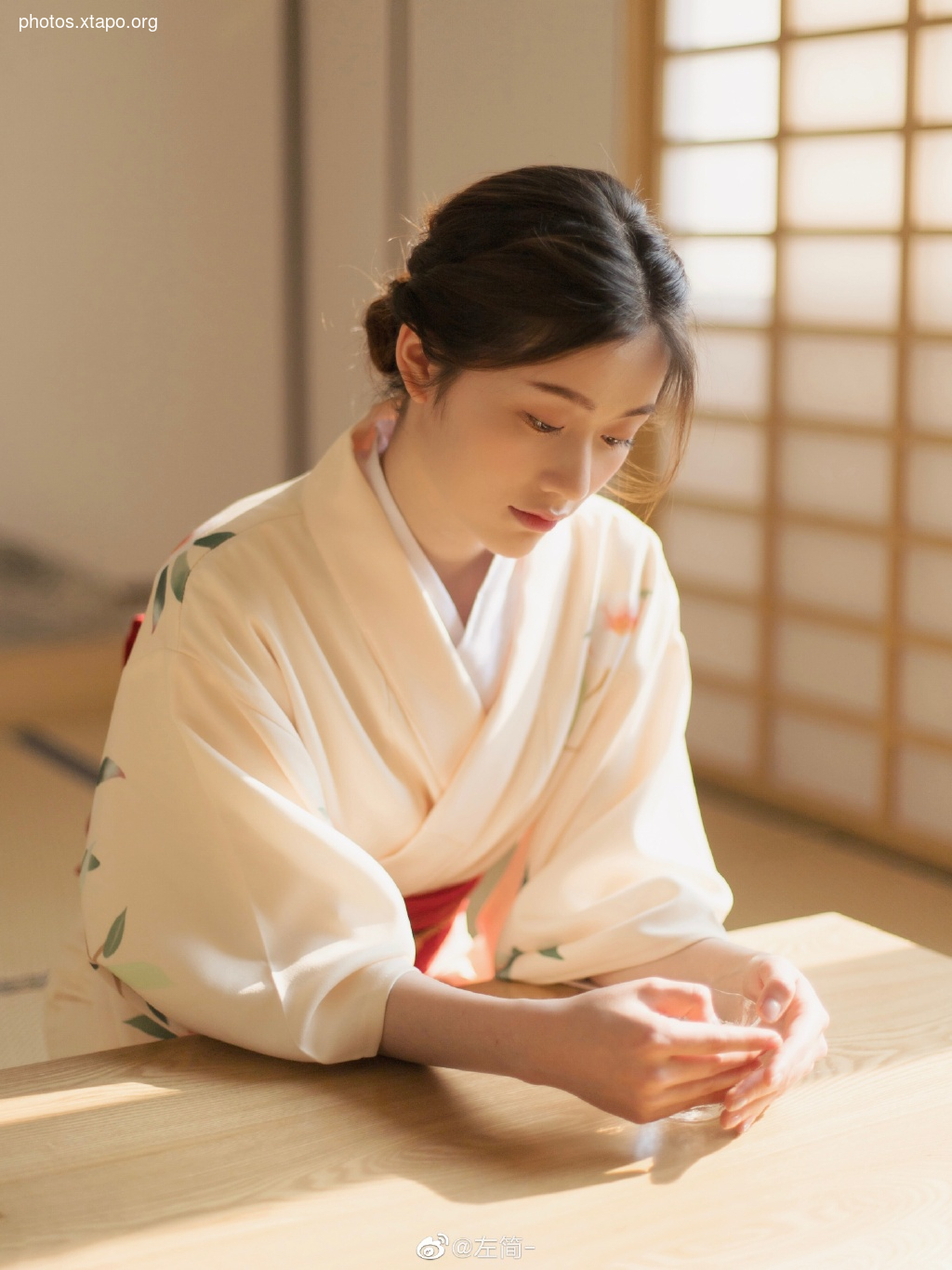 A young woman in a delicate, off-white kimono, adorned with subtle floral patterns, sits gracefully at a low table. Sunlight streams in, illuminating her serene expression and the intricate details of her attire. The soft, natural light and tranquil setting create a peaceful and elegant atmosphere.