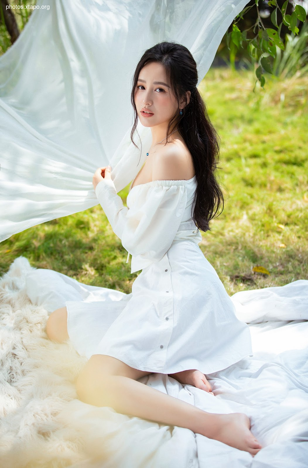 A young woman in a white off-the-shoulder dress sits on a white blanket outdoors.  She is posing for a photograph.