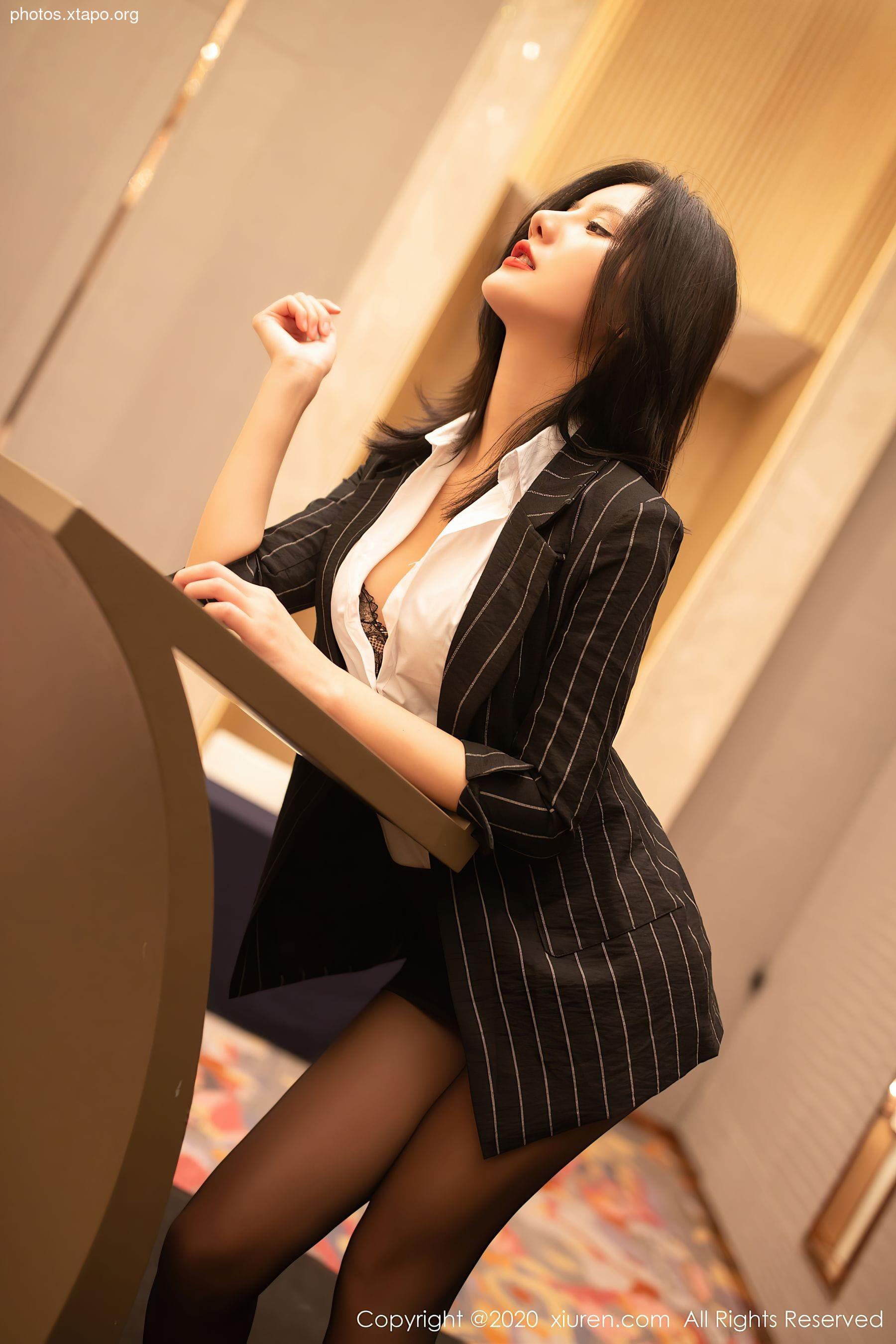 A woman in a pinstripe blazer and black stockings leans against a counter, exuding confidence and allure.