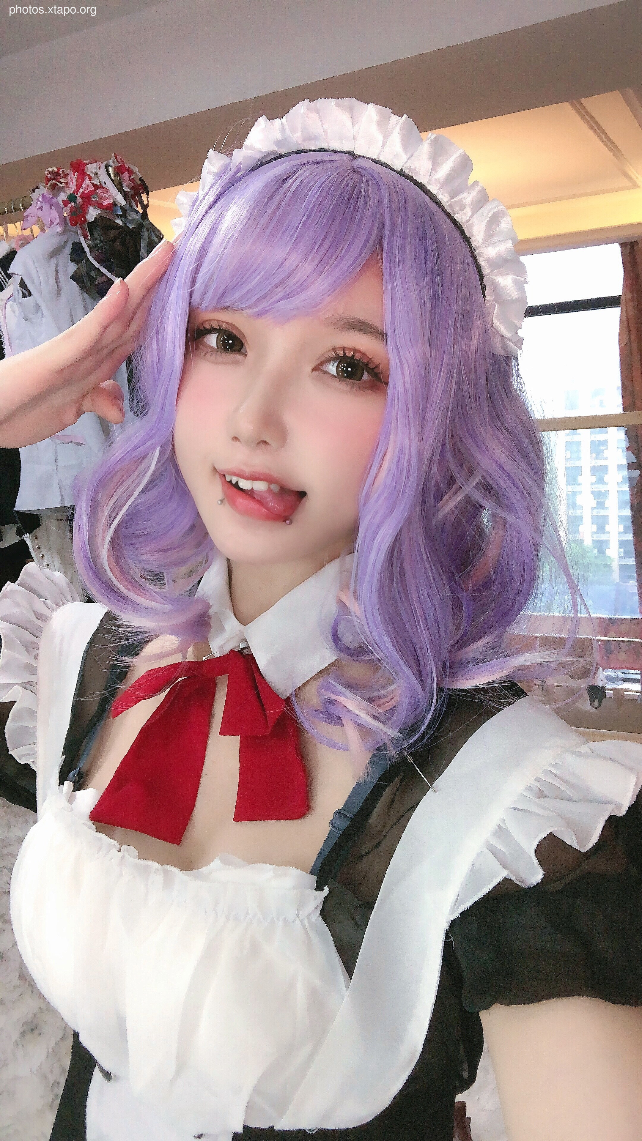 A cute girl in a maid cosplay with purple hair and a red bow.  She's giving a playful salute.