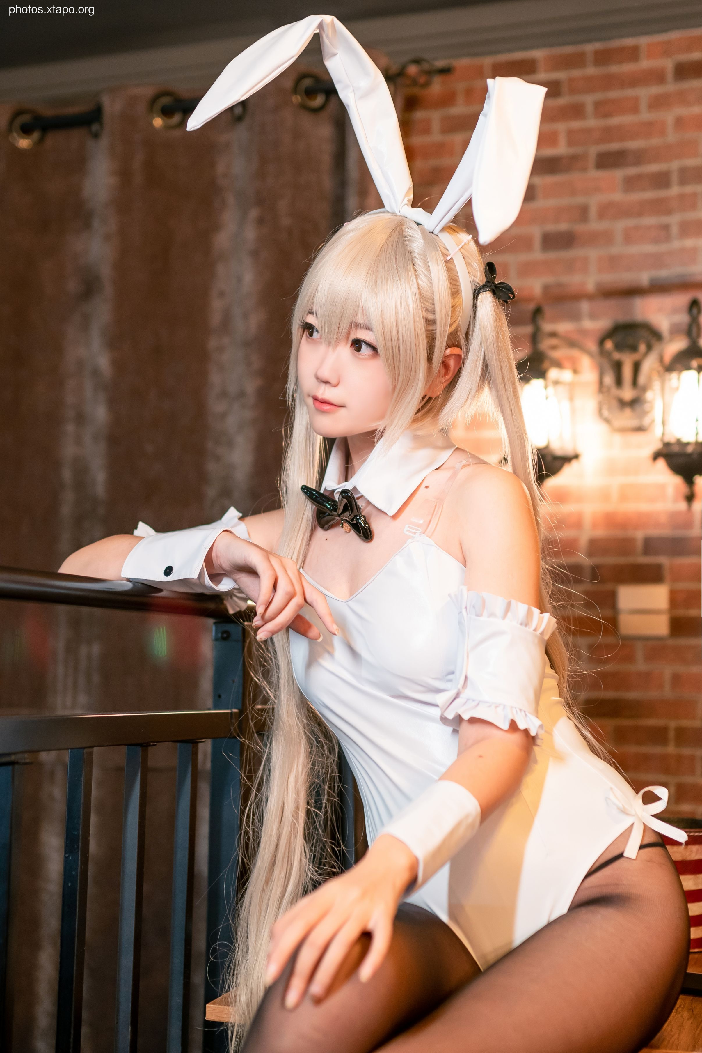 A young woman in a bunny girl costume sits on a bar rail, looking off to the side. She has long blonde hair and is wearing a white bunny outfit with black tights.