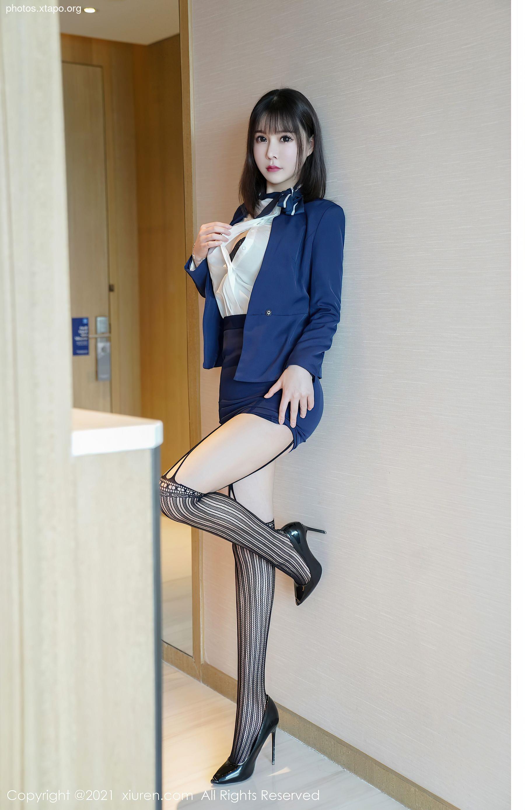 A woman in a navy blue blazer and white blouse, paired with a black and white striped stockings and black high heels. She is standing against a light beige wall.