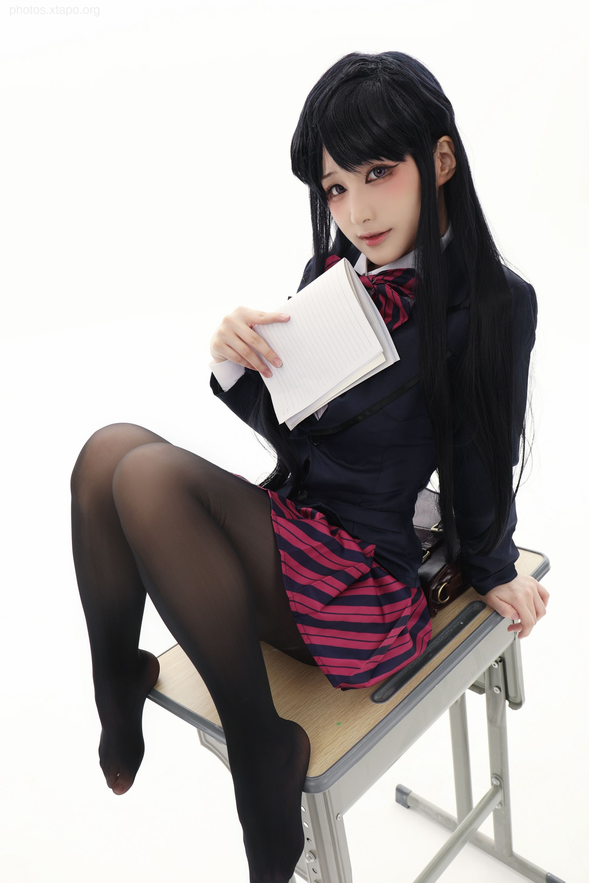A young woman in schoolgirl cosplay sits on a desk, holding a notebook.  She's dressed in a dark blazer, striped skirt, and black tights.