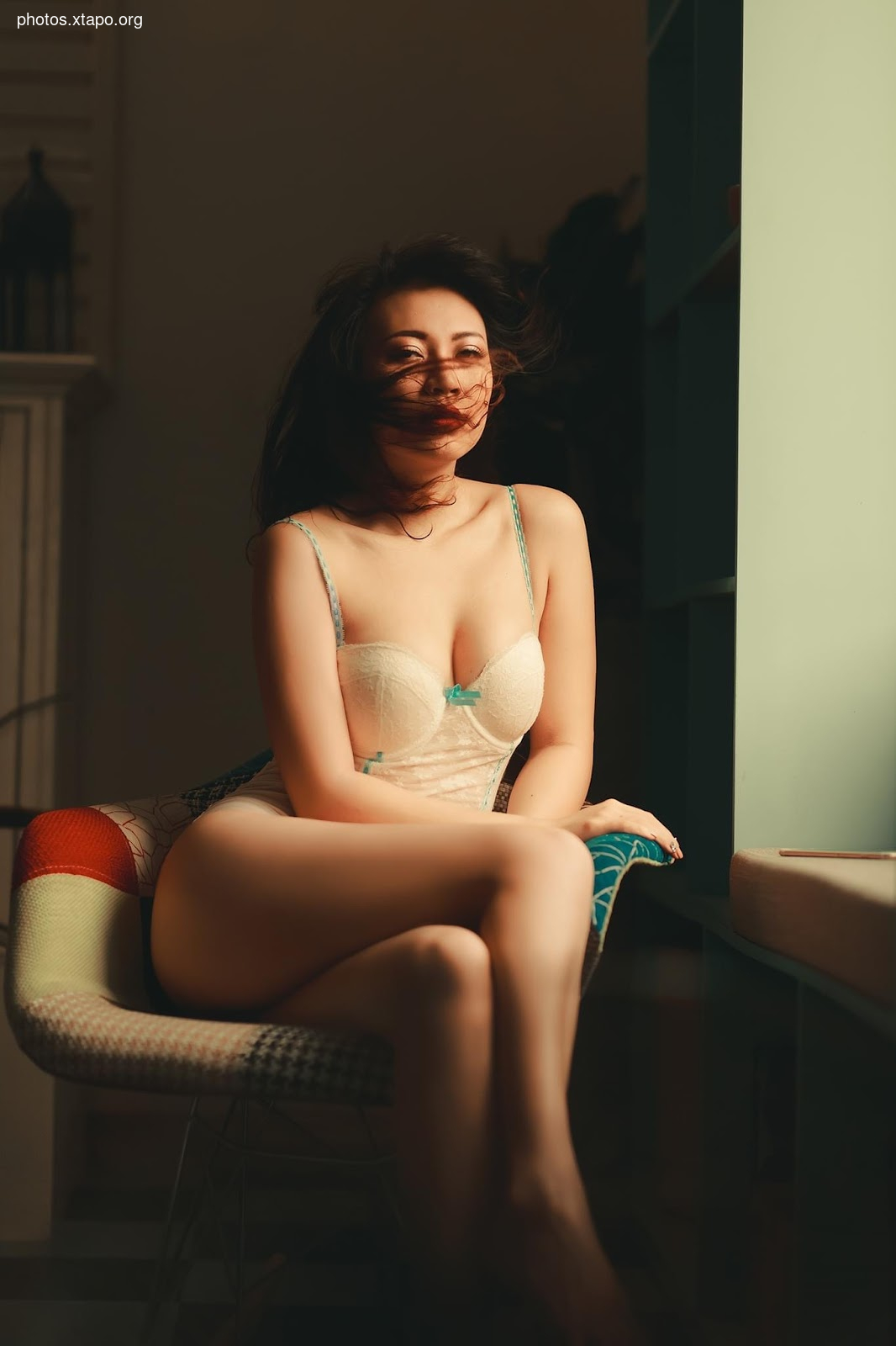 A woman sits on a chair, wearing a white lace lingerie. Her hair is blowing in the wind.