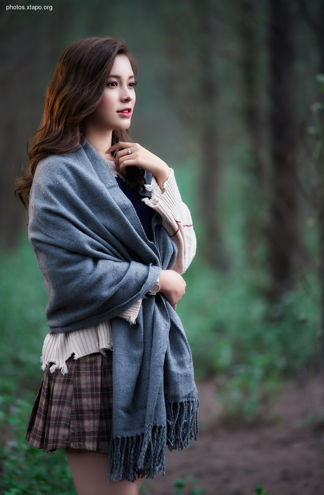 A young woman with long brown hair stands in a forest, wrapped in a gray shawl. She gazes thoughtfully into the distance.