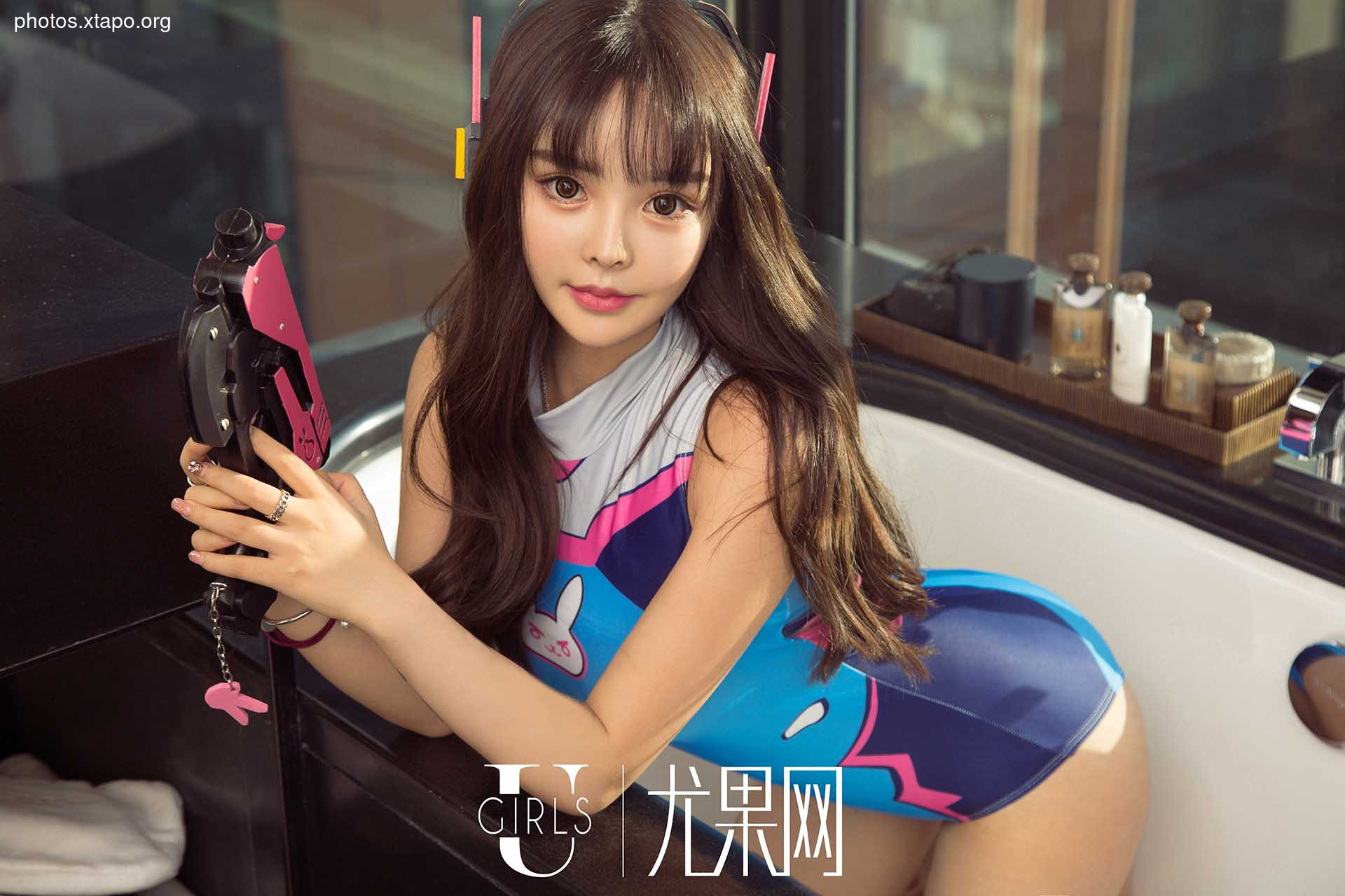 A young woman poses in a D.Va cosplay costume, holding a pink prop weapon. She is sitting in a bathtub, surrounded by bathroom amenities.