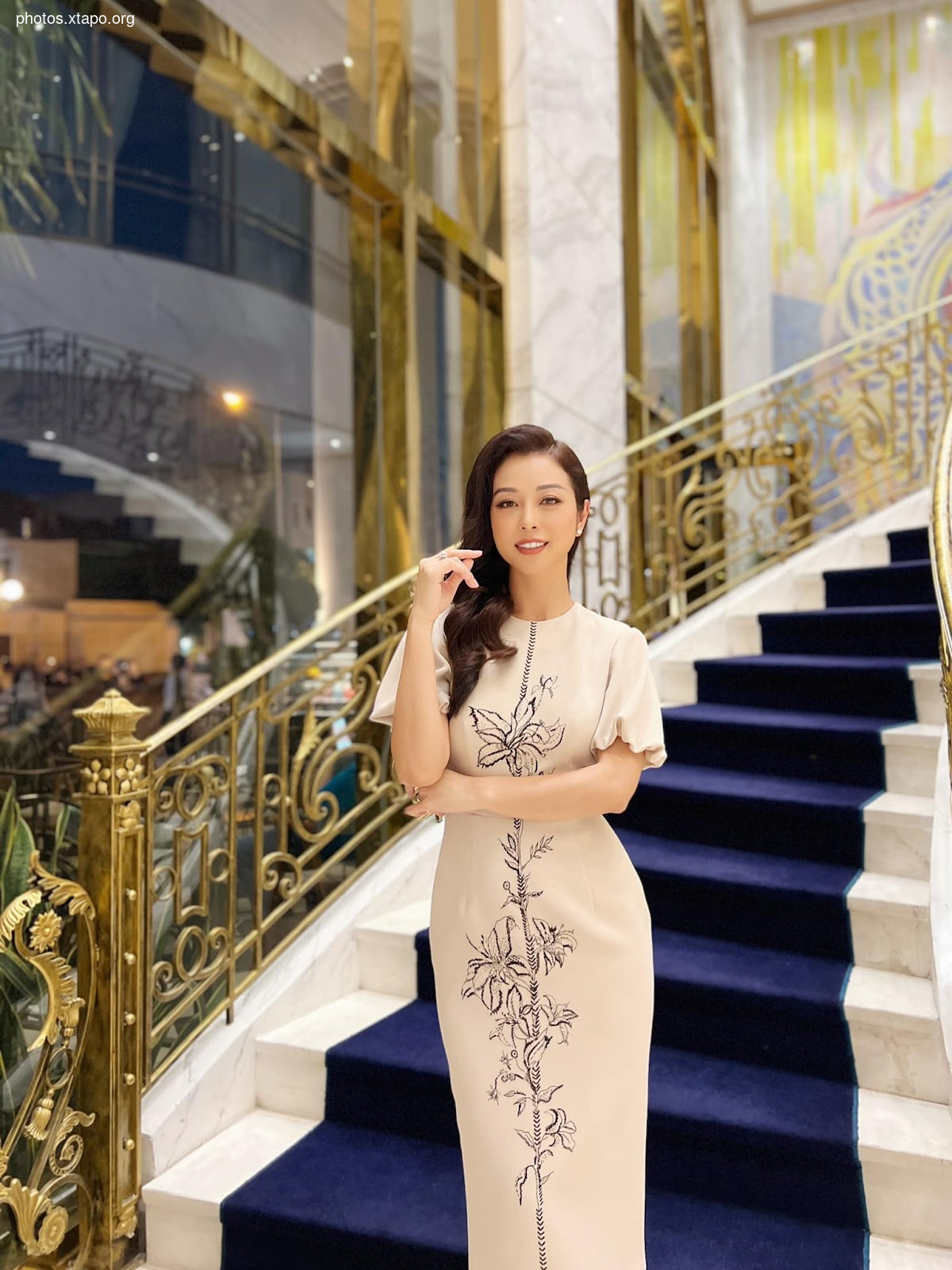 A woman in a beige, floor-length gown adorned with a delicate floral design stands on a grand staircase. The dress features a modest neckline and short sleeves, complementing her sophisticated style. The setting is luxurious, with a marble staircase and ornate gold railings.