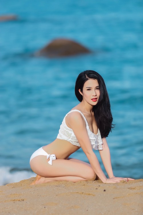A woman in a white bikini poses on a sandy beach with the ocean in the background.