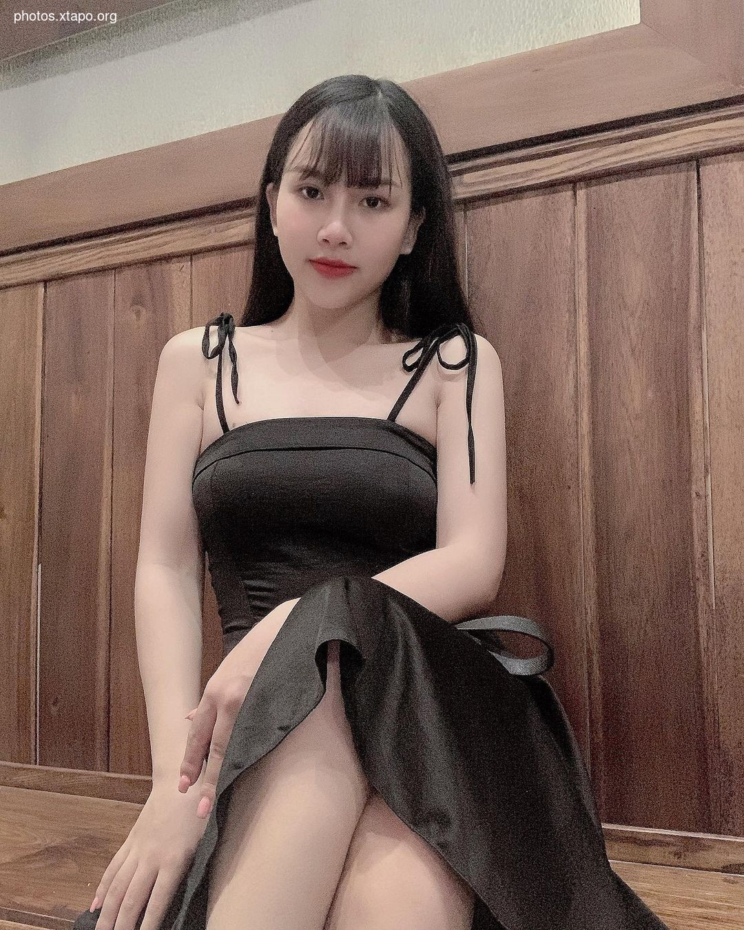 A young woman poses in a stylish black dress with thin straps against a wooden background.