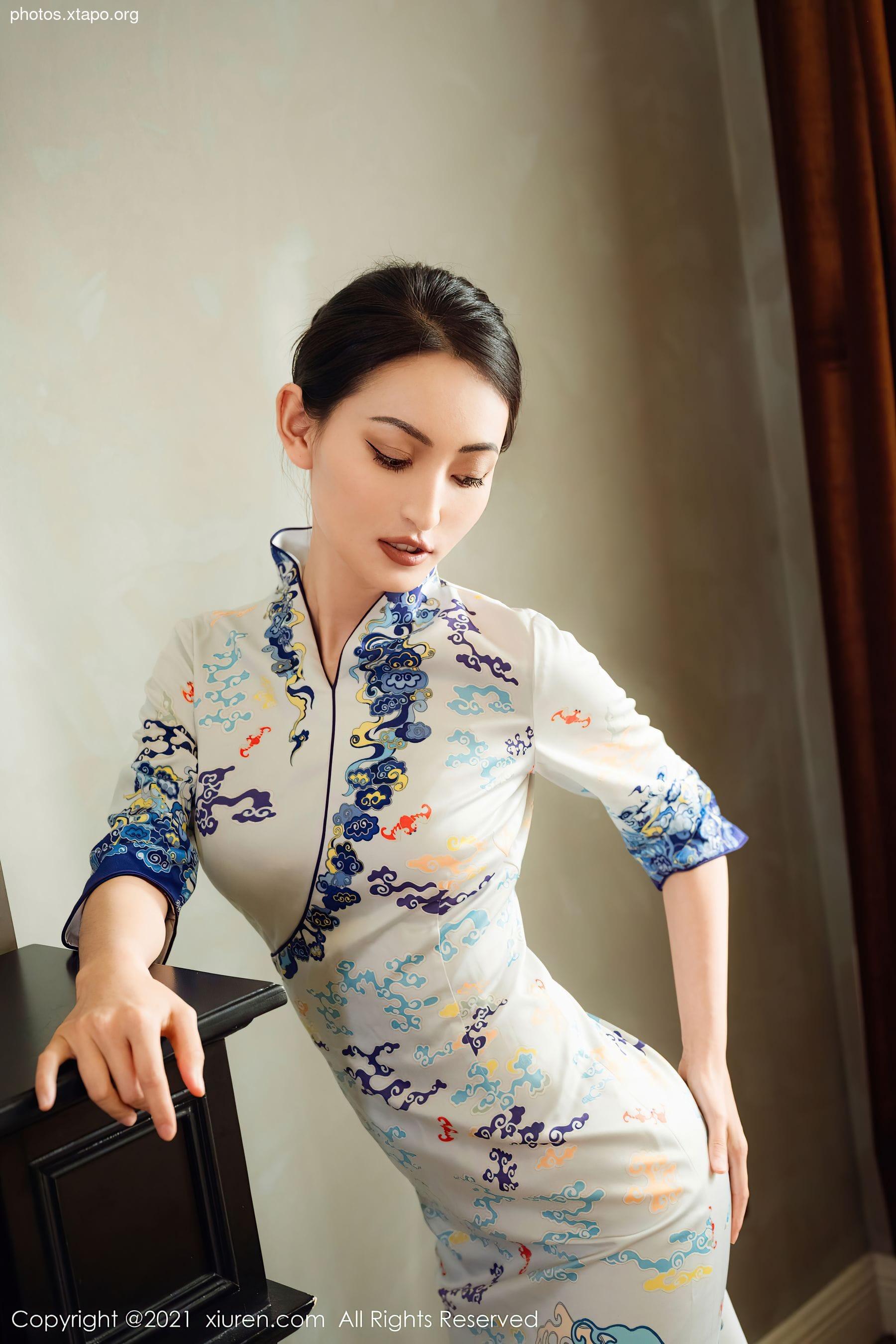 A young woman elegantly poses in a traditional Chinese cheongsam dress. The dress features a delicate floral pattern and intricate detailing. The woman's pose and expression convey grace and poise.