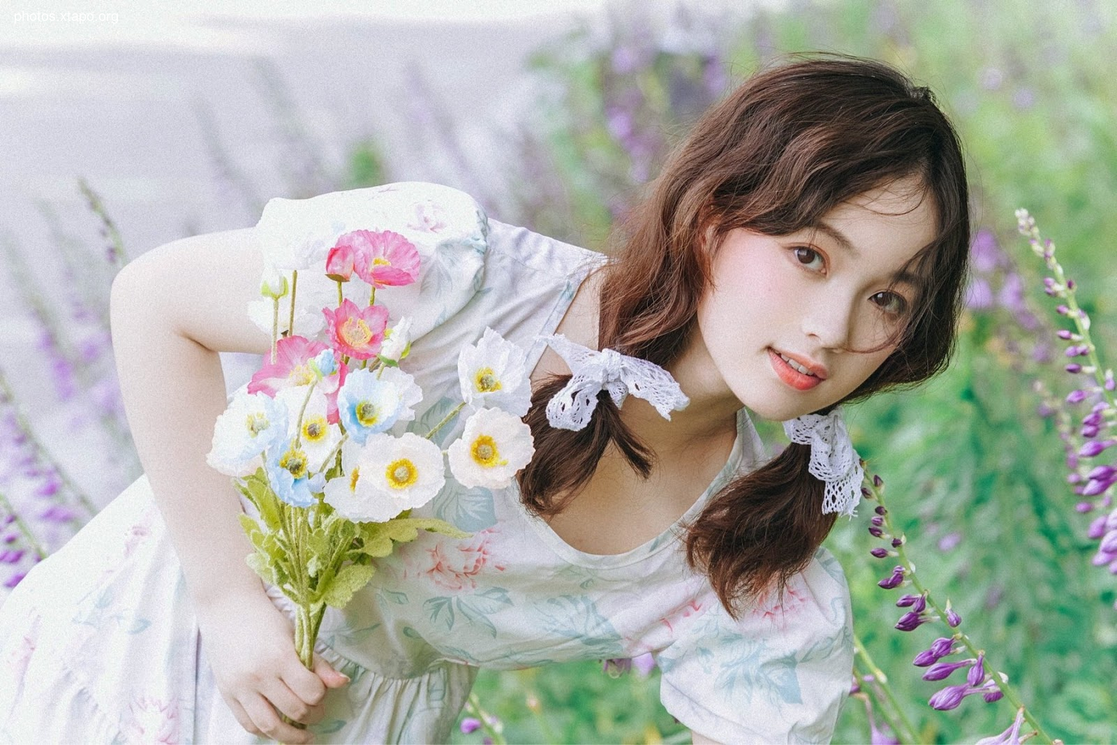 A young woman in a floral dress smiles gently as she holds a bouquet of colorful poppies. The image is soft and dreamy, with a focus on the woman's gentle beauty and the vibrant flowers surrounding her.