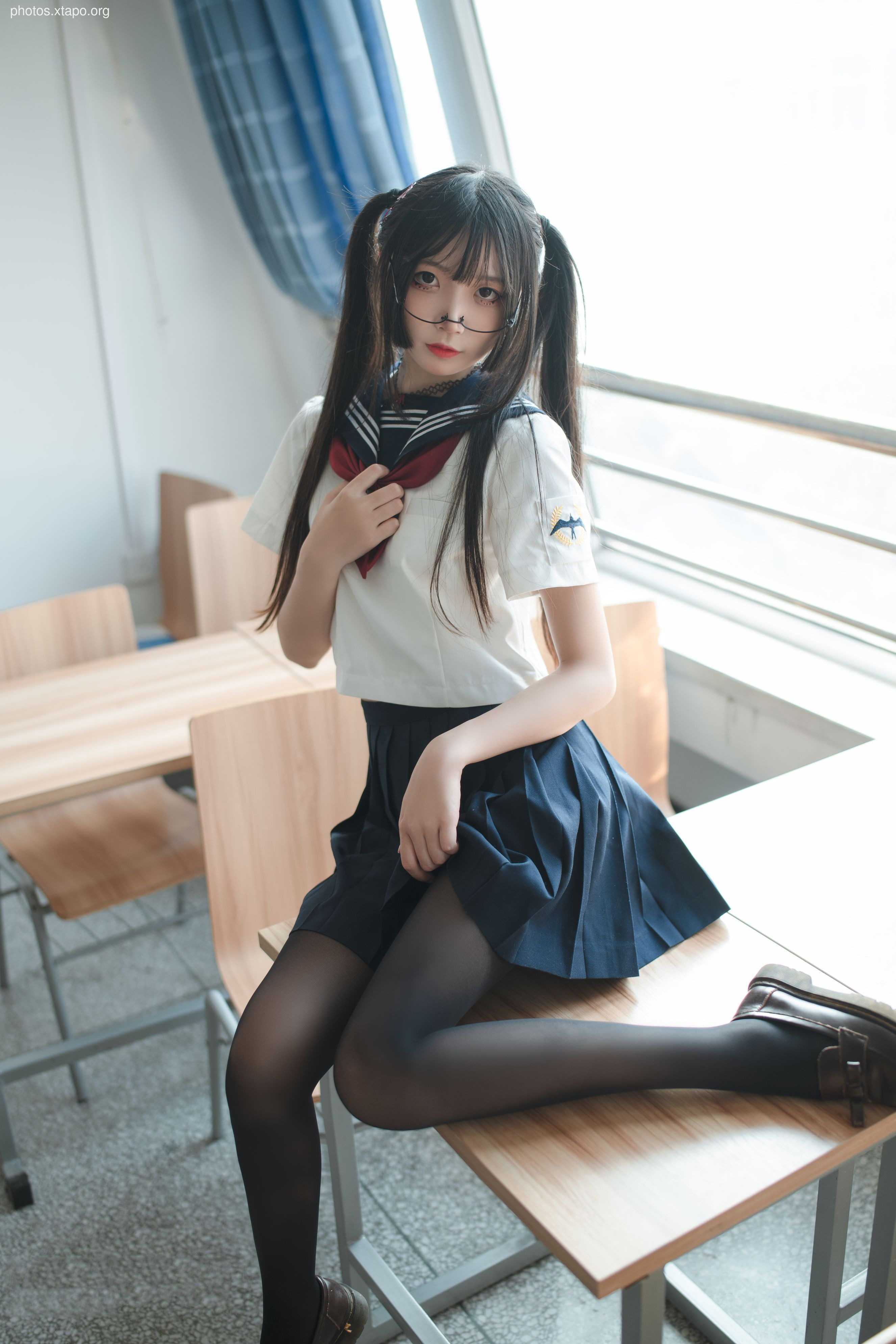 A young woman in a Japanese schoolgirl sailor uniform sits at a desk, wearing black tights and glasses.