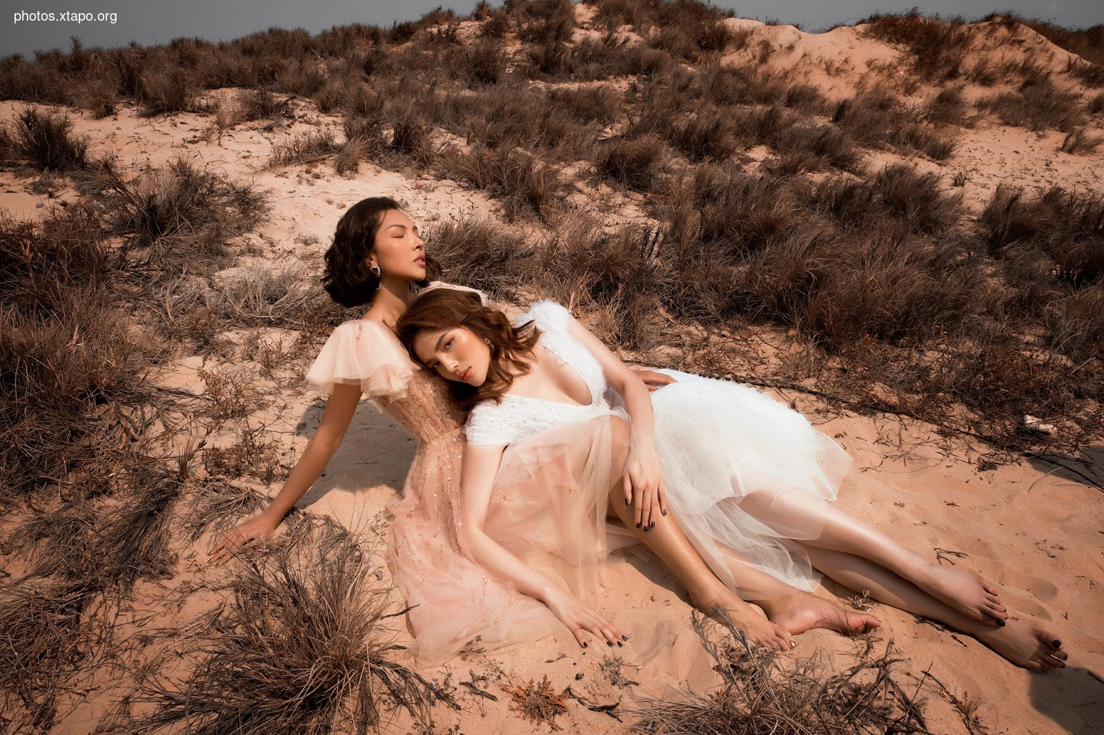 Two women in flowing gowns embrace amidst the serene beauty of a sandy landscape.