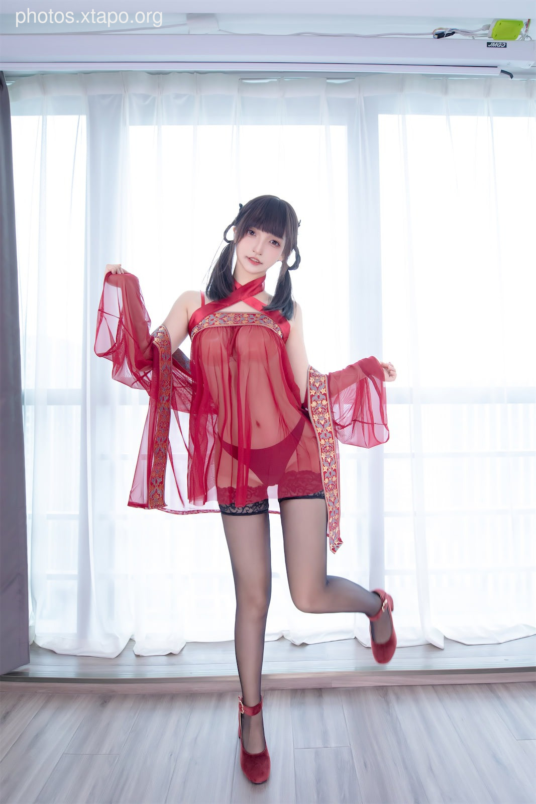 A young woman in a beautiful red sheer cheongsam poses elegantly near a window.