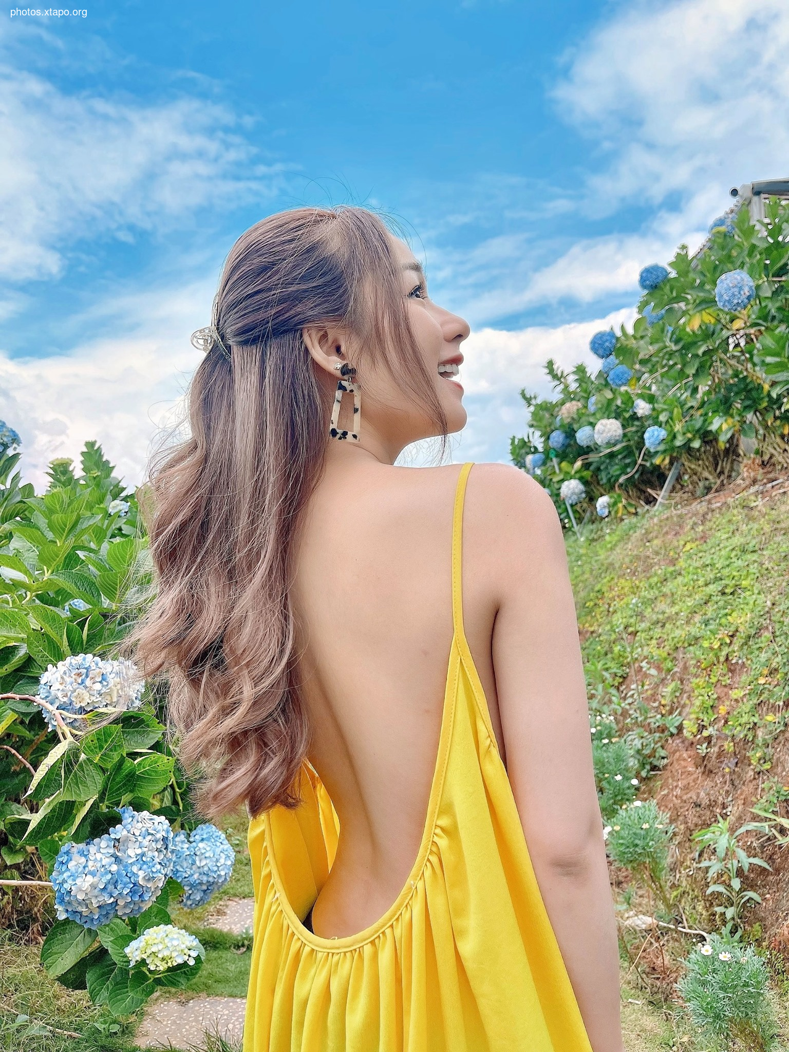 Woman in a backless yellow dress enjoying a day in a flower garden.  The sky is blue and the flowers are vibrant.