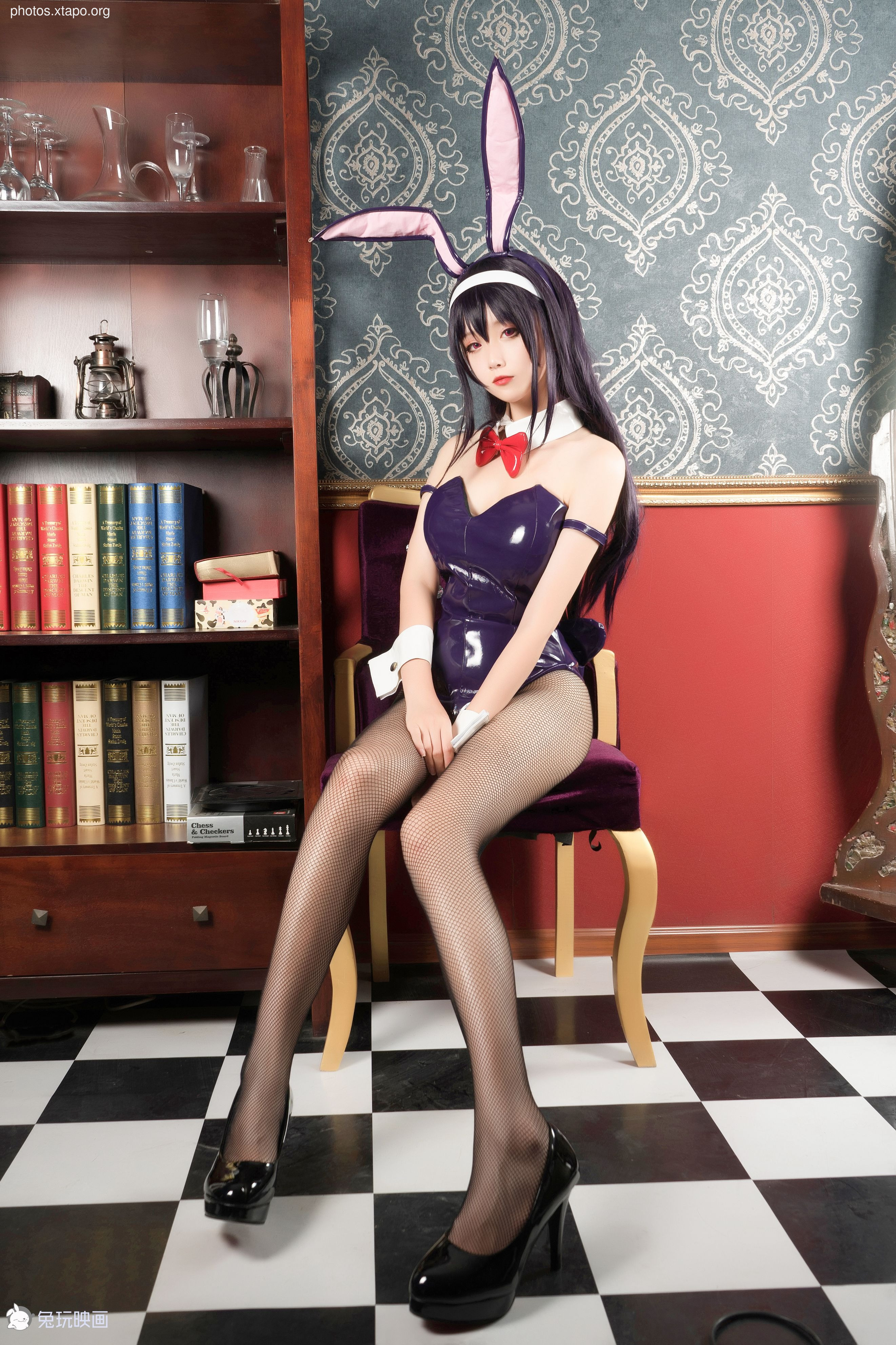 A young woman in a purple bunny girl costume sits on a chair in a library.  She is wearing black fishnet stockings and black high heels.