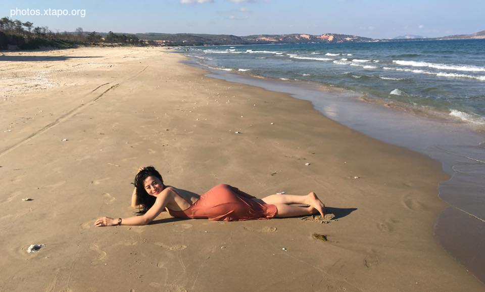 A woman in a peach-colored dress relaxes on a sandy beach near the ocean.