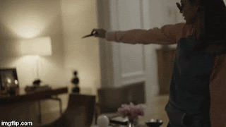 A woman in a denim jacket holds scissors, extending her arm towards an unseen target in a dimly lit room.