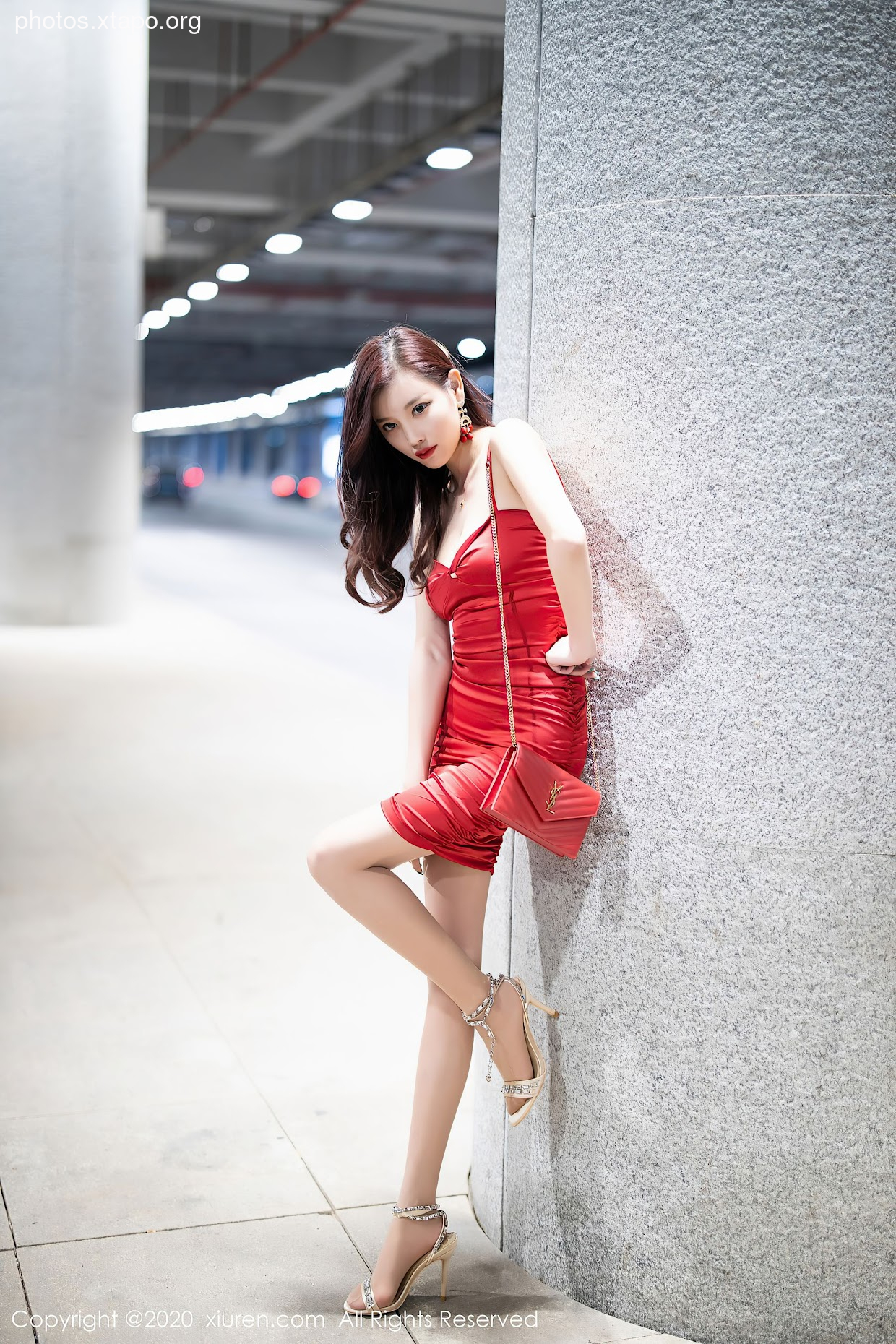 A woman in a red dress poses against a pillar. She carries a handbag and wears high heels.