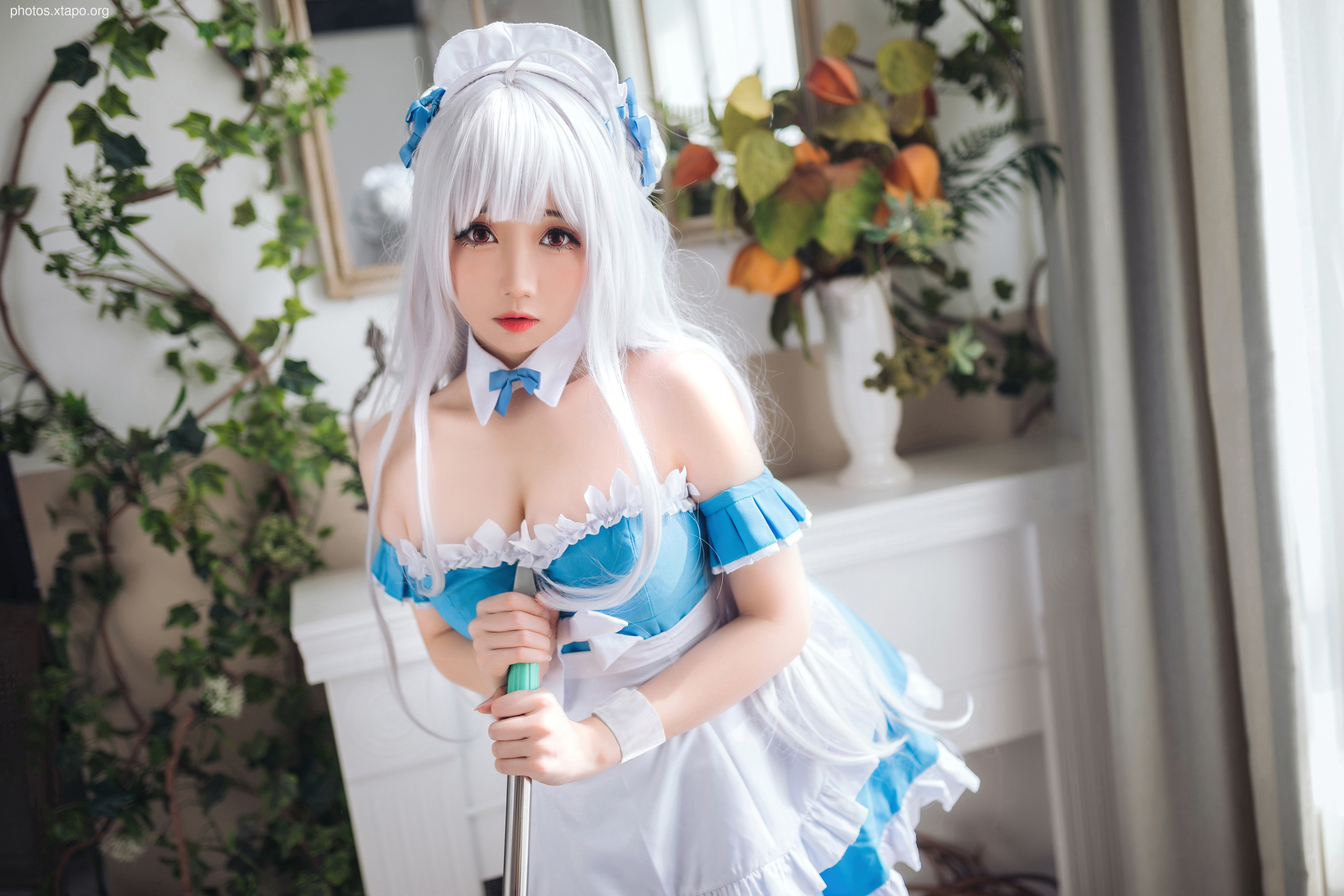 A young woman cosplaying as a maid, holding a mop. She is wearing a white and blue maid outfit with a white apron and a white headband. She has long white hair and is looking at the camera.