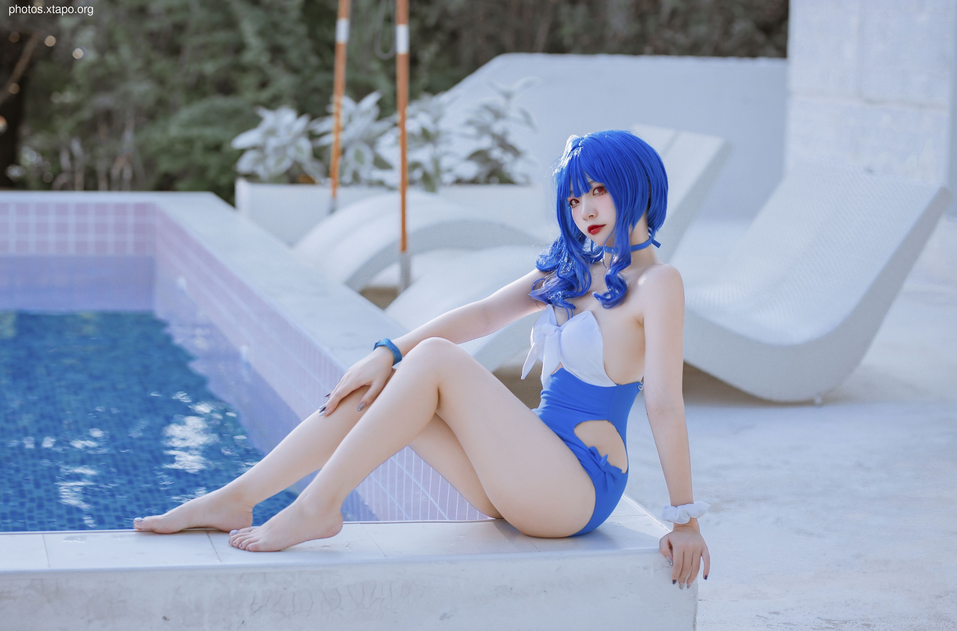 A young woman in a blue and white swimsuit poses by a pool.  She has long blue hair and is wearing makeup. The setting is luxurious and modern.