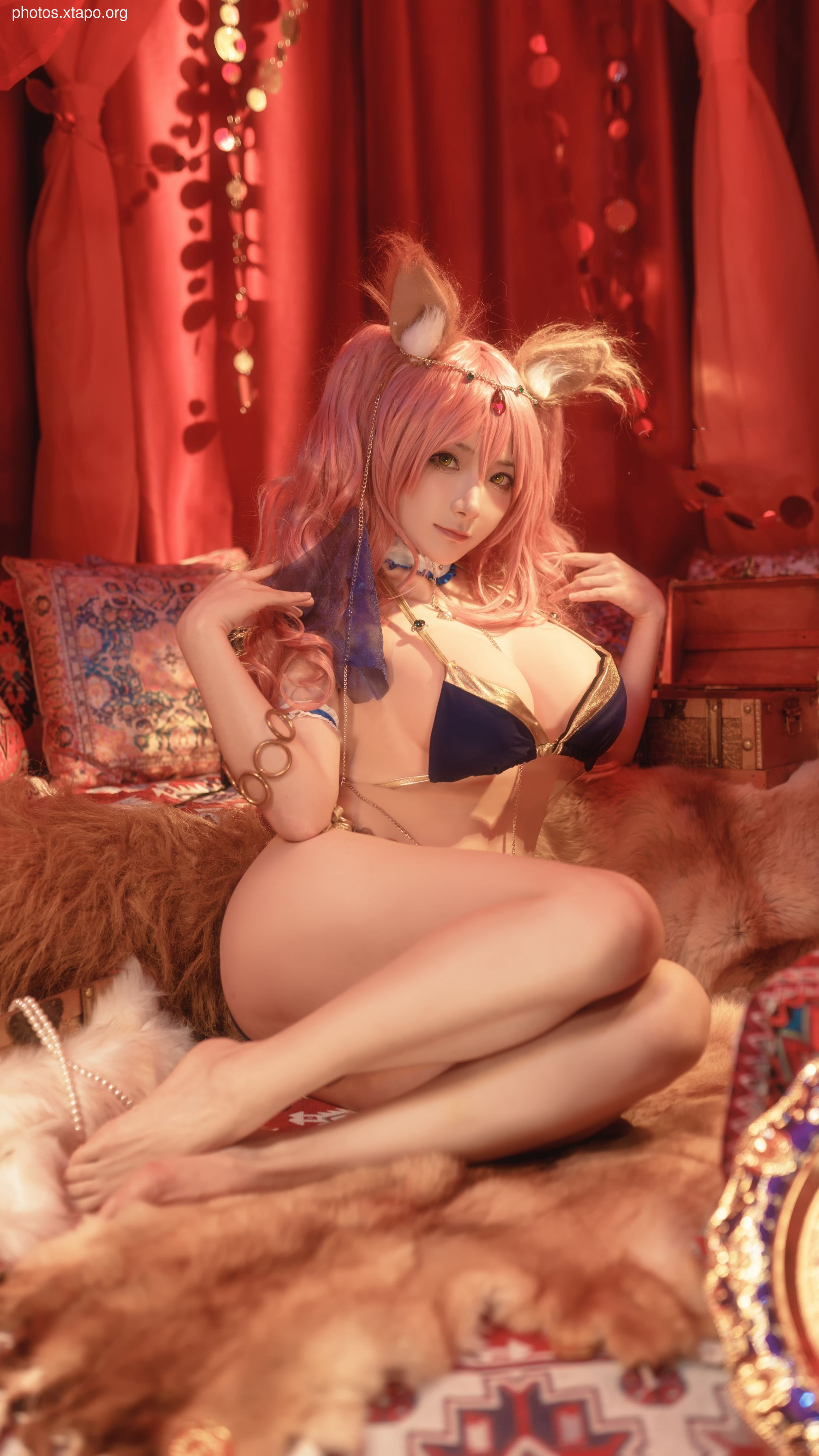 A stunning cosplay of a female character in a fantasy setting.  She's wearing a revealing, gold and blue bikini-style outfit with fox ears and pink hair.