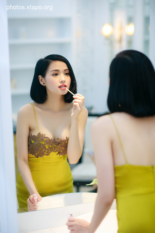 A young woman in a yellow dress applies lip gloss while looking in a mirror.