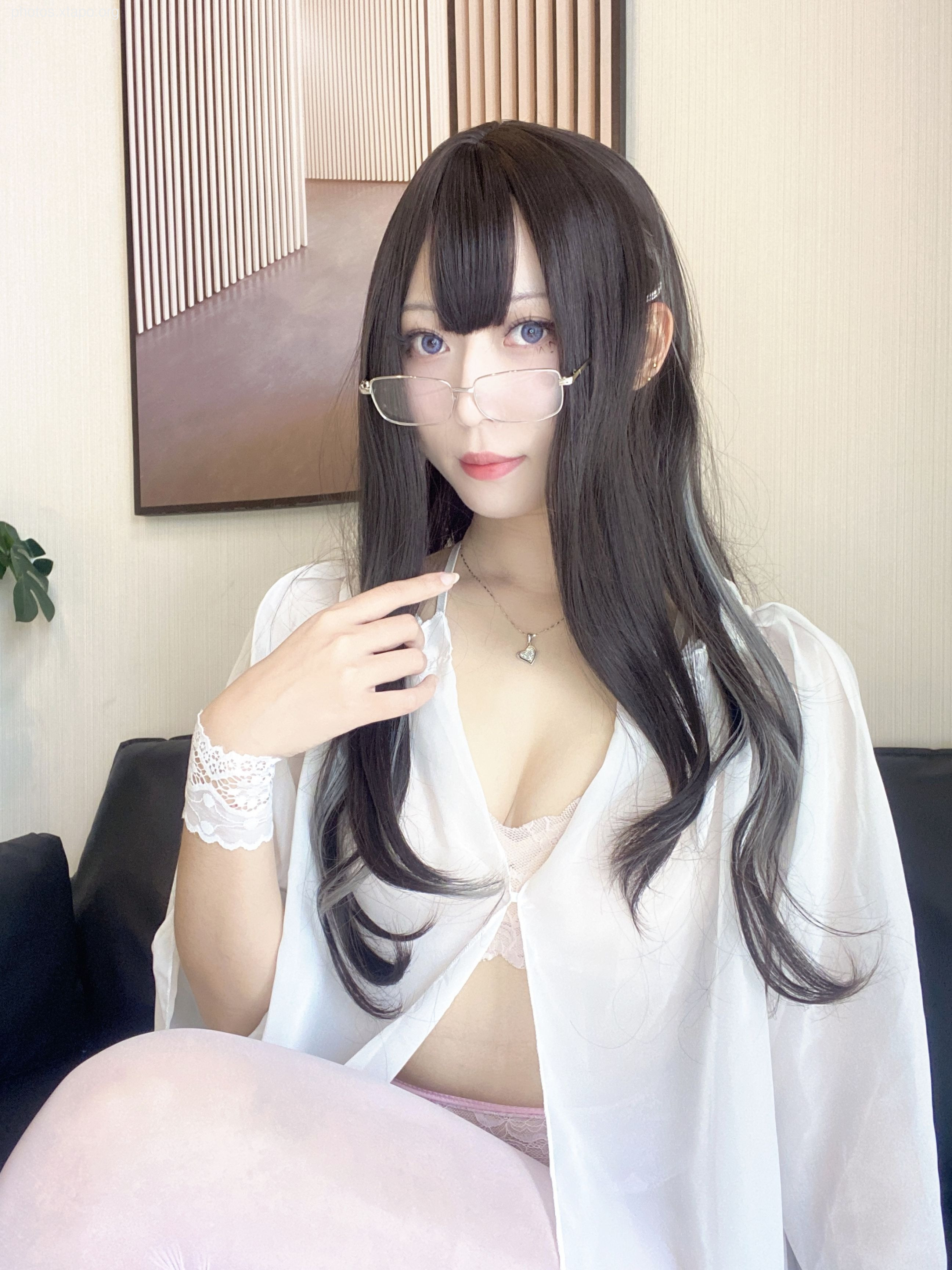 A young woman with long dark hair, wearing glasses and a white shirt, playfully looks at the camera. She is sitting on a couch and is wearing light pink tights.