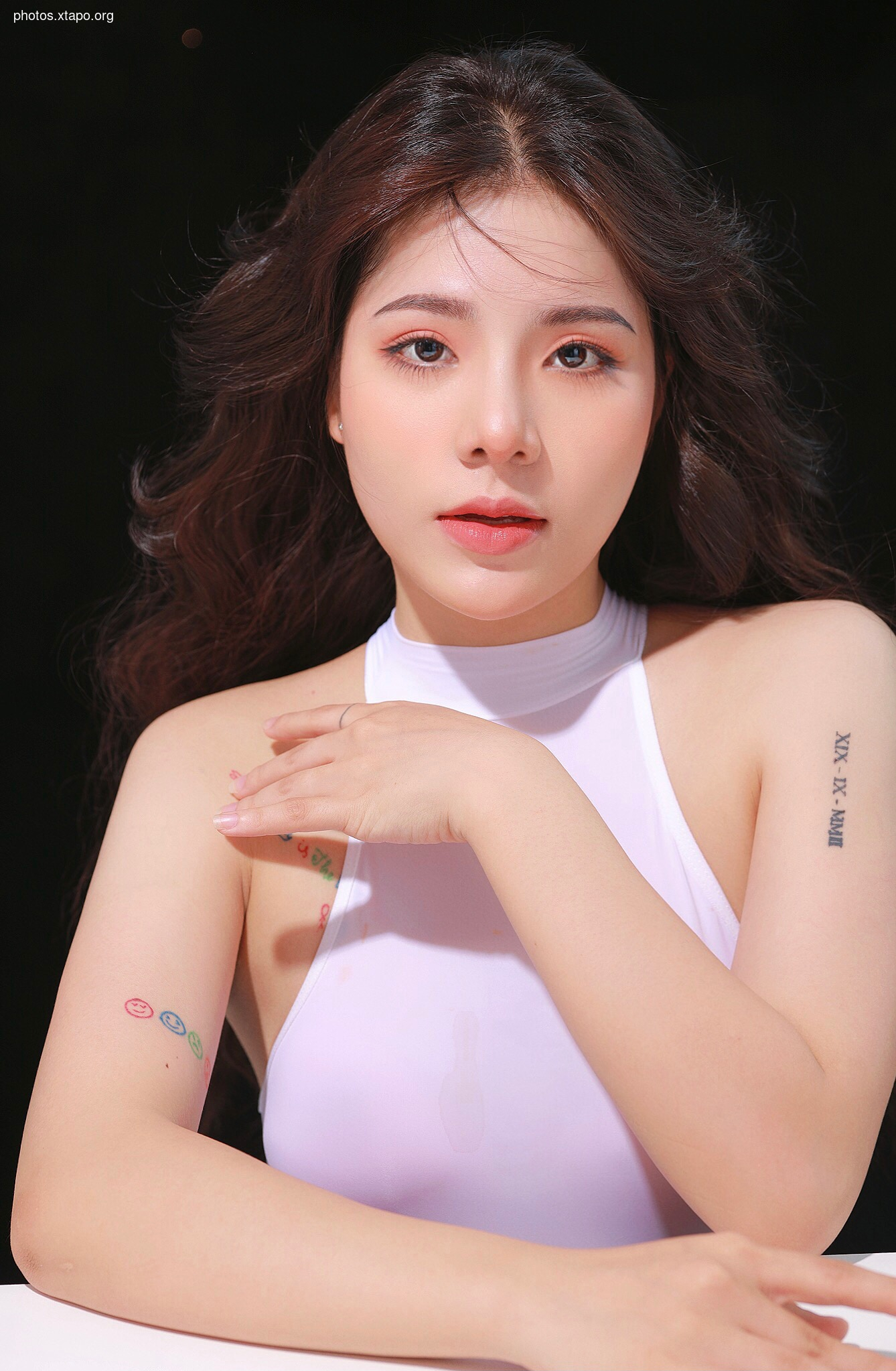 A young woman with long brown hair poses in a white tank top. She has delicate features and subtle tattoos on her arms.