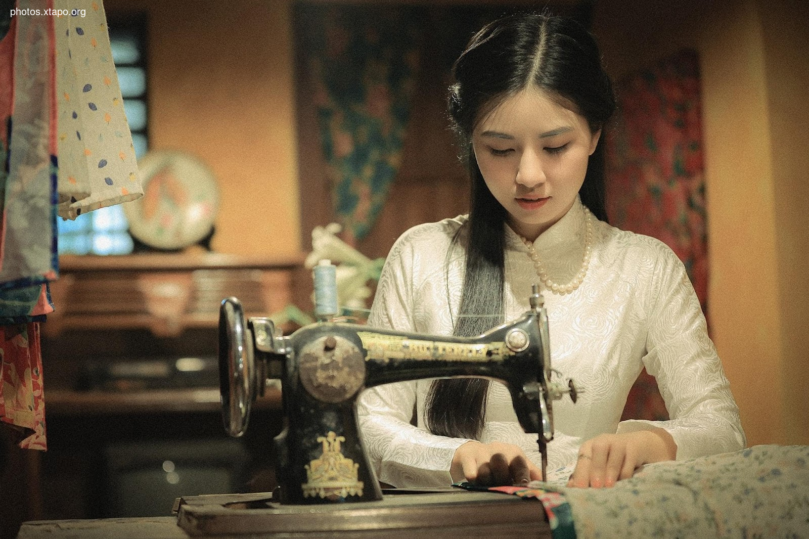 A young woman in traditional Vietnamese clothing sits at an antique sewing machine, meticulously working on a piece of fabric. The scene is set in a rustic, vintage-style room, adding to the nostalgic charm of the image.