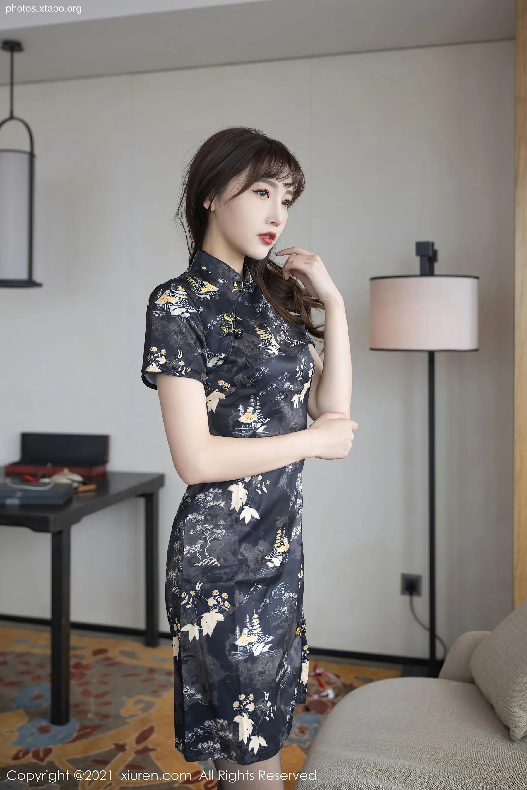 A young woman models a stylish dark-colored cheongsam with a floral print. The dress features a mandarin collar and short sleeves. She is posing in a well-lit room with a modern, Asian-inspired decor.