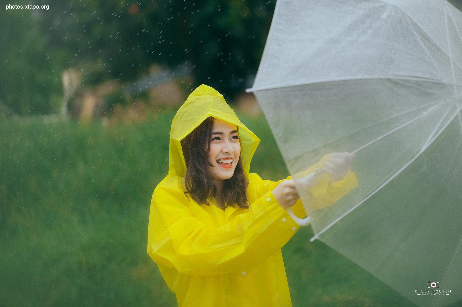 A young woman smiles brightly while walking in the rain, using a clear umbrella to stay dry. She's wearing a cheerful yellow raincoat.