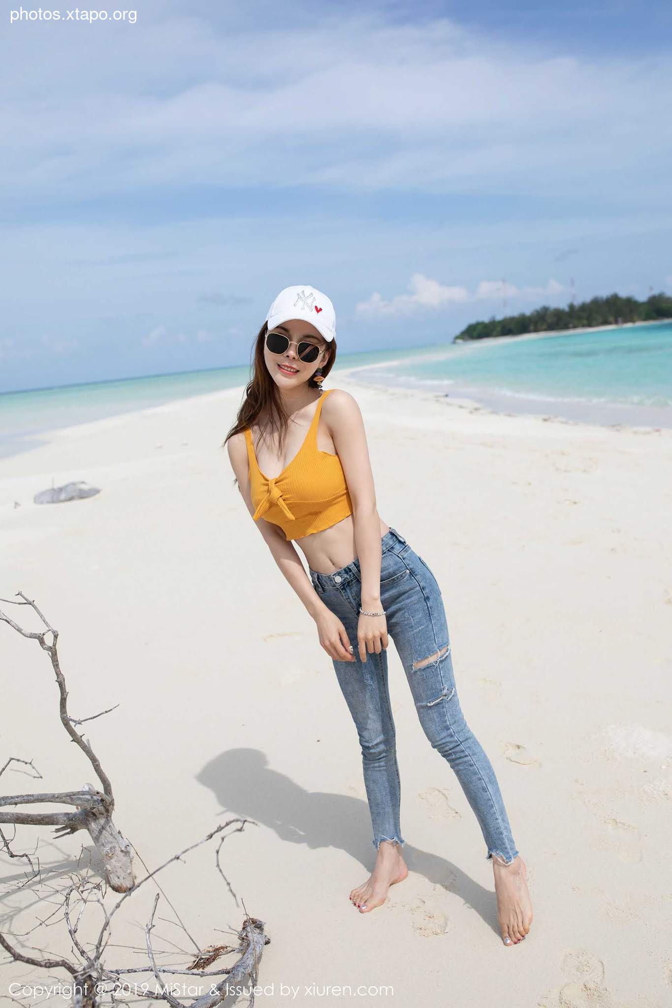 A young woman in a yellow top and ripped jeans poses on a tropical beach.