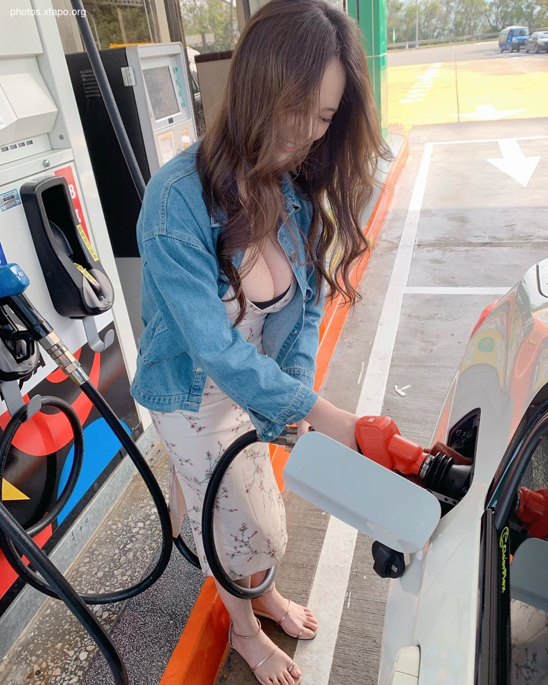 A woman in a denim jacket and floral dress pumps gas into her car at a gas station.
