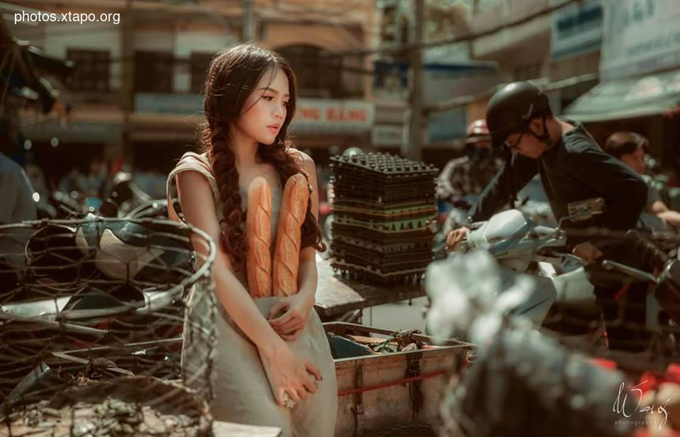 A young Asian woman with long brown hair holds two baguettes while standing in a bustling market. She is wearing a light beige dress and appears pensive. The background shows a vibrant street scene with motorbikes and other people.