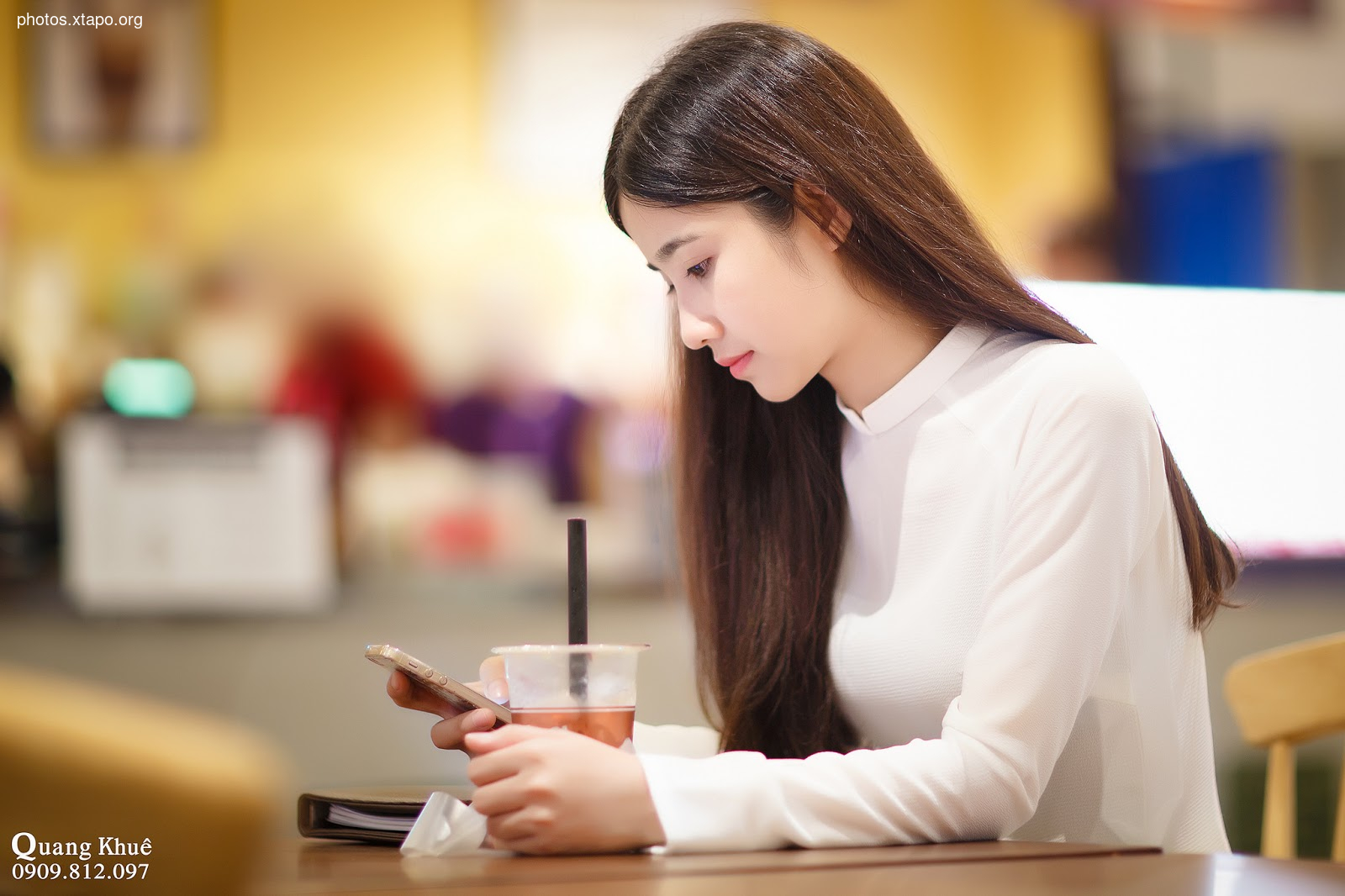 A young woman sits at a table in a cafe, using her smartphone and enjoying a drink.