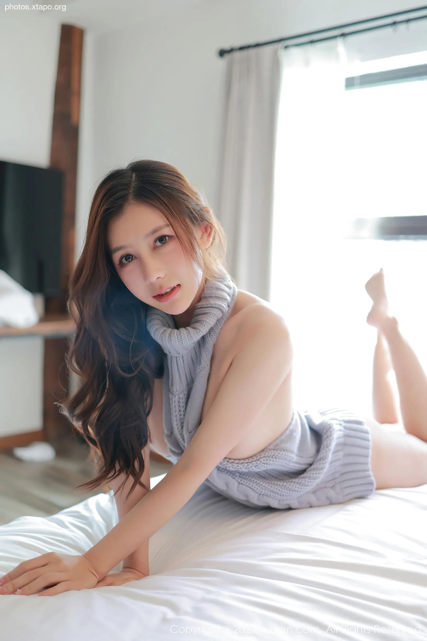 A young woman with long brown hair is lying on a white bed wearing a light gray turtleneck sweater.  She is looking at the camera.