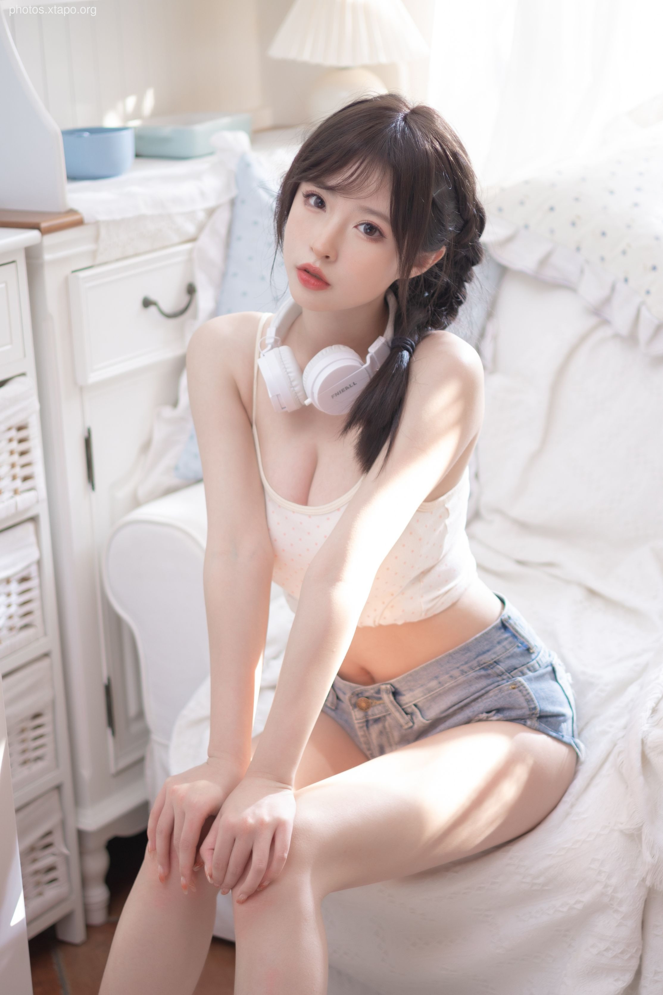 A young woman with long dark hair sits on a white couch wearing a white cropped tank top and denim shorts. She has white headphones around her neck and is looking at the camera.