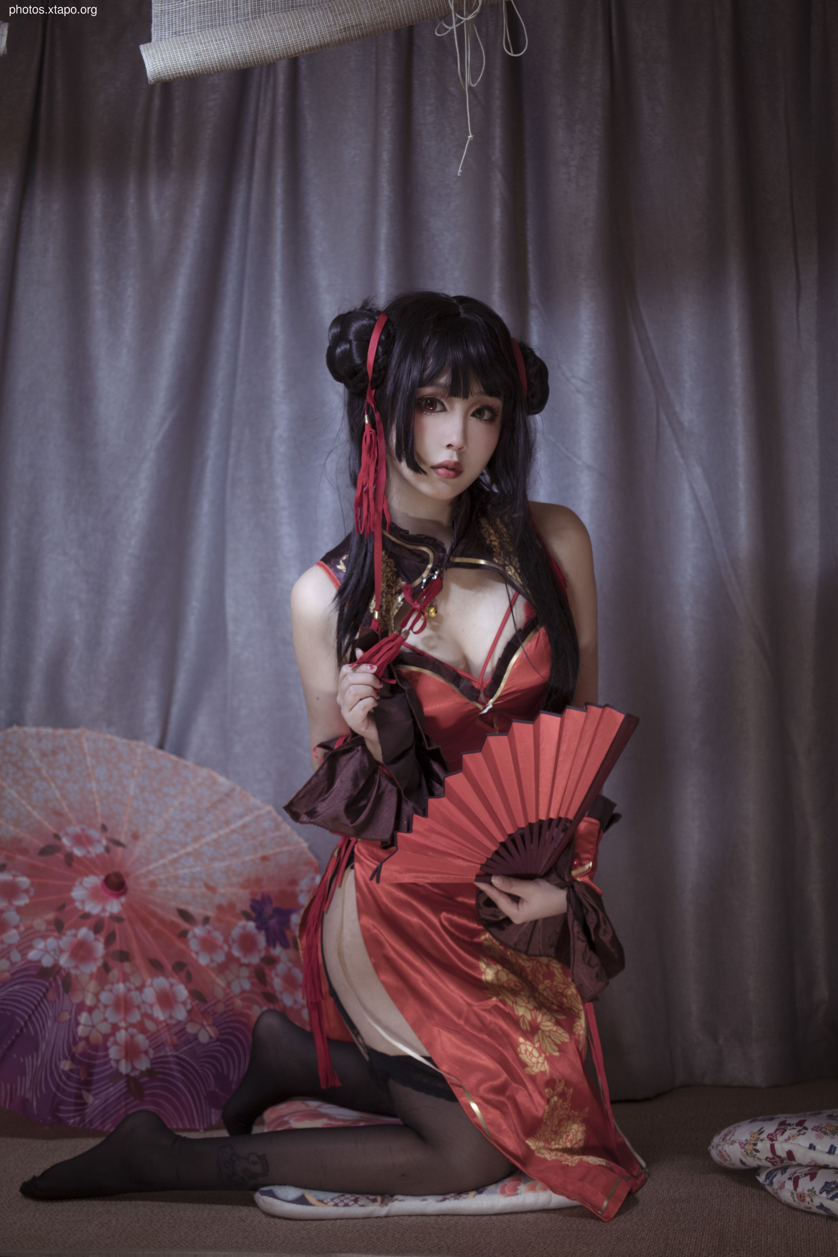 A woman in cosplay, dressed in a vibrant red and gold traditional Chinese qipao, holds a red hand fan.  She is posed elegantly against a muted grey background.