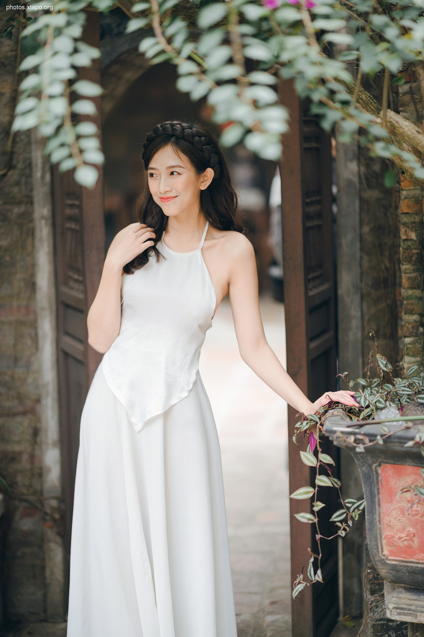 A young woman in a flowing white halter neck dress stands gracefully in a garden setting.  She has dark hair styled in a braid and is positioned near an ornate doorway.