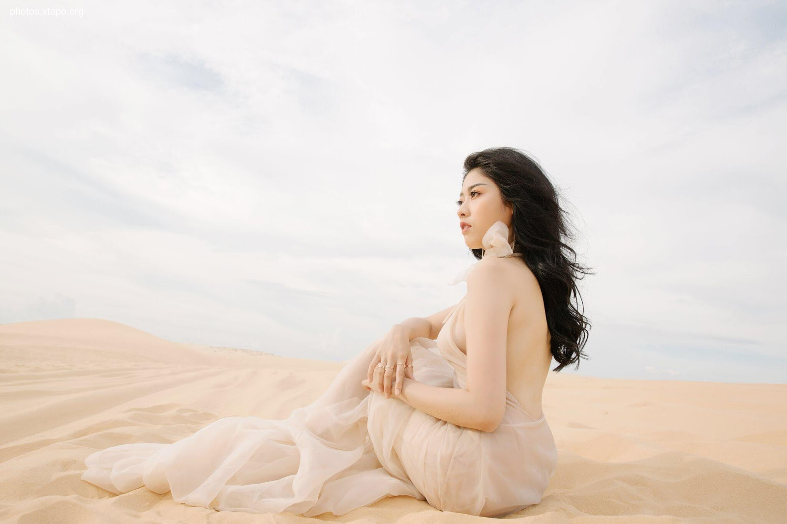 A woman in a flowing beige gown sits in a desert, gazing into the distance. The vast expanse of sand dunes and the clear sky create a serene and ethereal atmosphere.