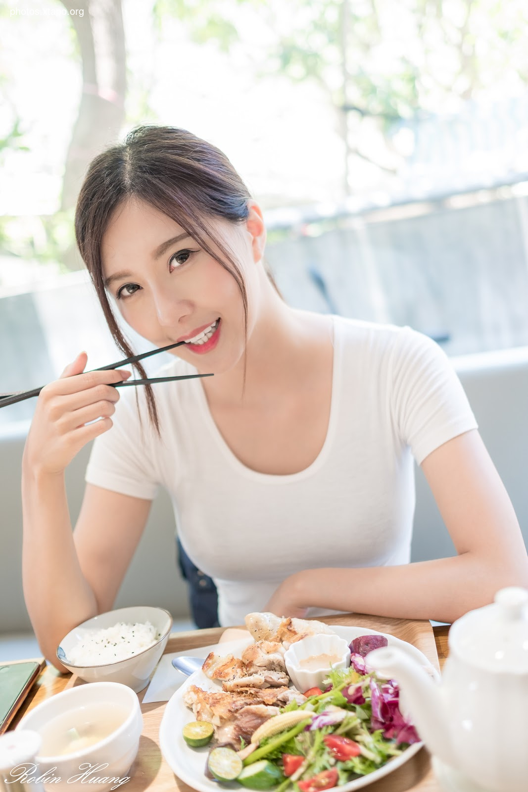 A young woman smiles while enjoying a healthy and delicious meal.  She is using chopsticks to eat a plate of grilled chicken, salad, and rice.