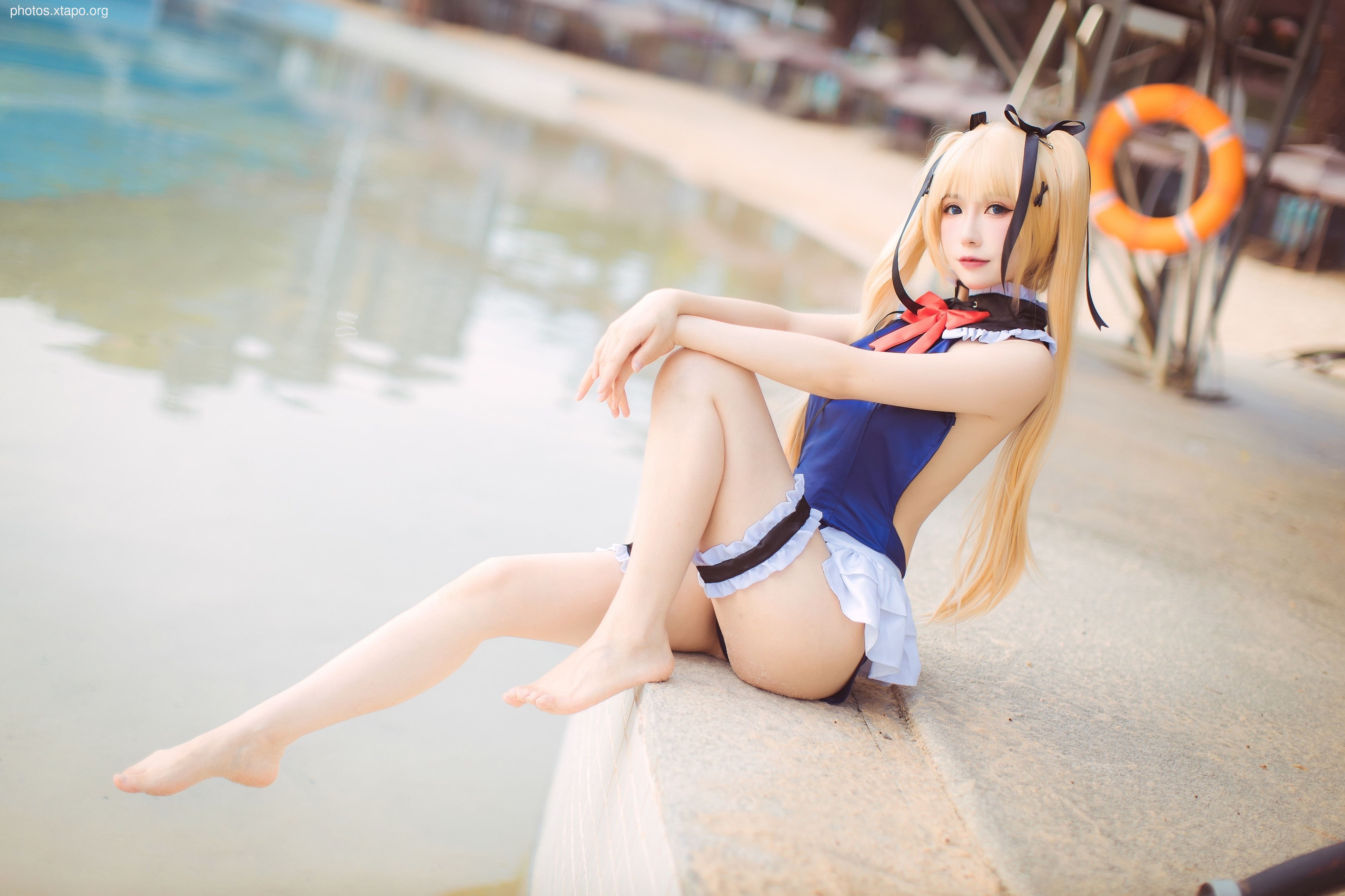A young woman in a blue and white swimsuit cosplay sits by a pool.  She has long blonde hair styled with ribbons.