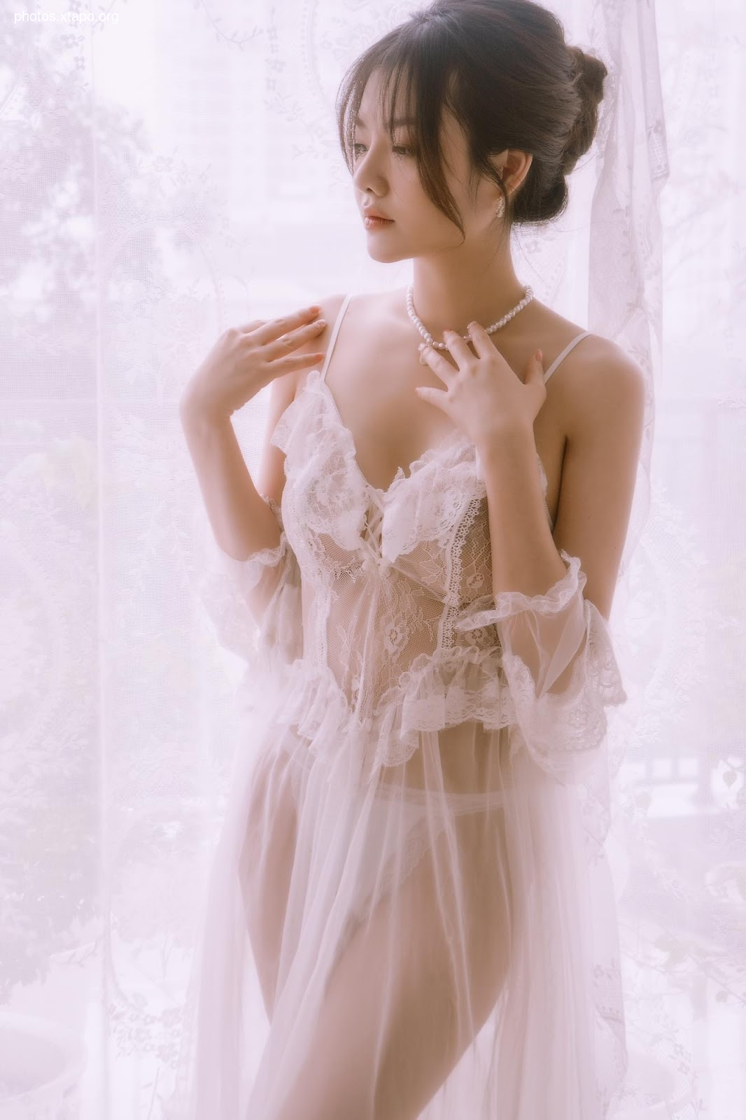 A woman in a delicate white lace lingerie dress stands by a window.