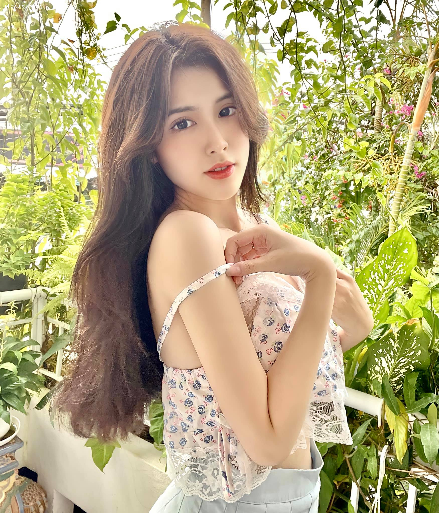 A young woman with long brown hair poses outdoors wearing a floral top and light blue pants.