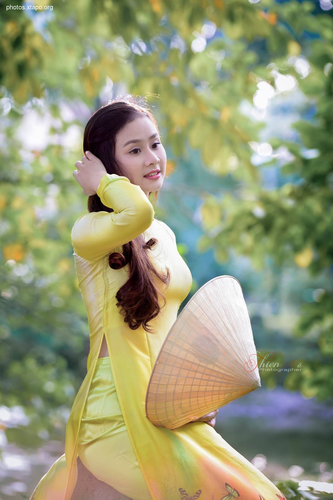 A young woman in a traditional Vietnamese Ao Dai dress poses outdoors with a conical hat.