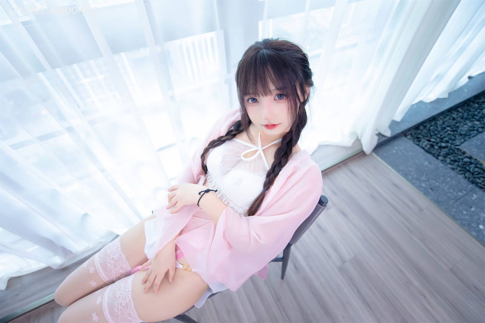 A young woman in a pink robe and white lingerie sits on a chair, looking directly at the camera.  She is near a window with white curtains.