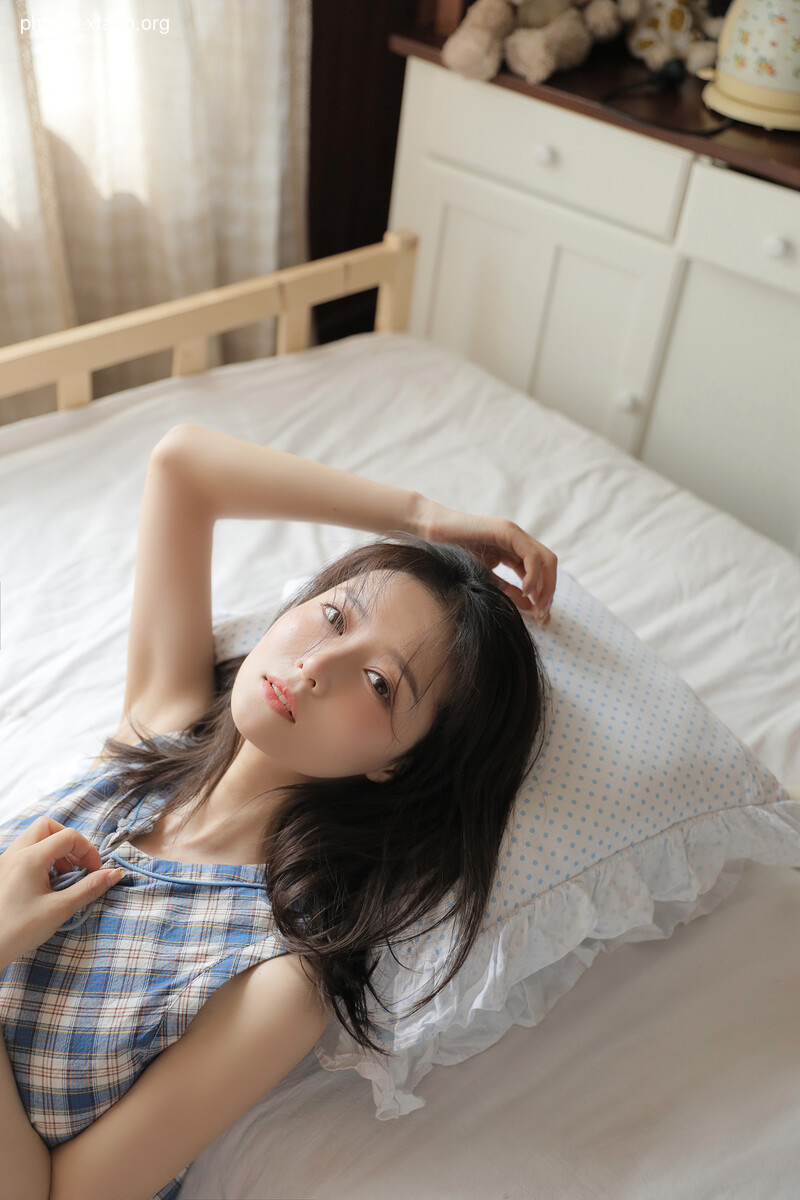 A young woman with long black hair relaxes on a bed, wearing a blue and white plaid sleeveless top. Soft lighting and a calm atmosphere enhance the image's serene mood.